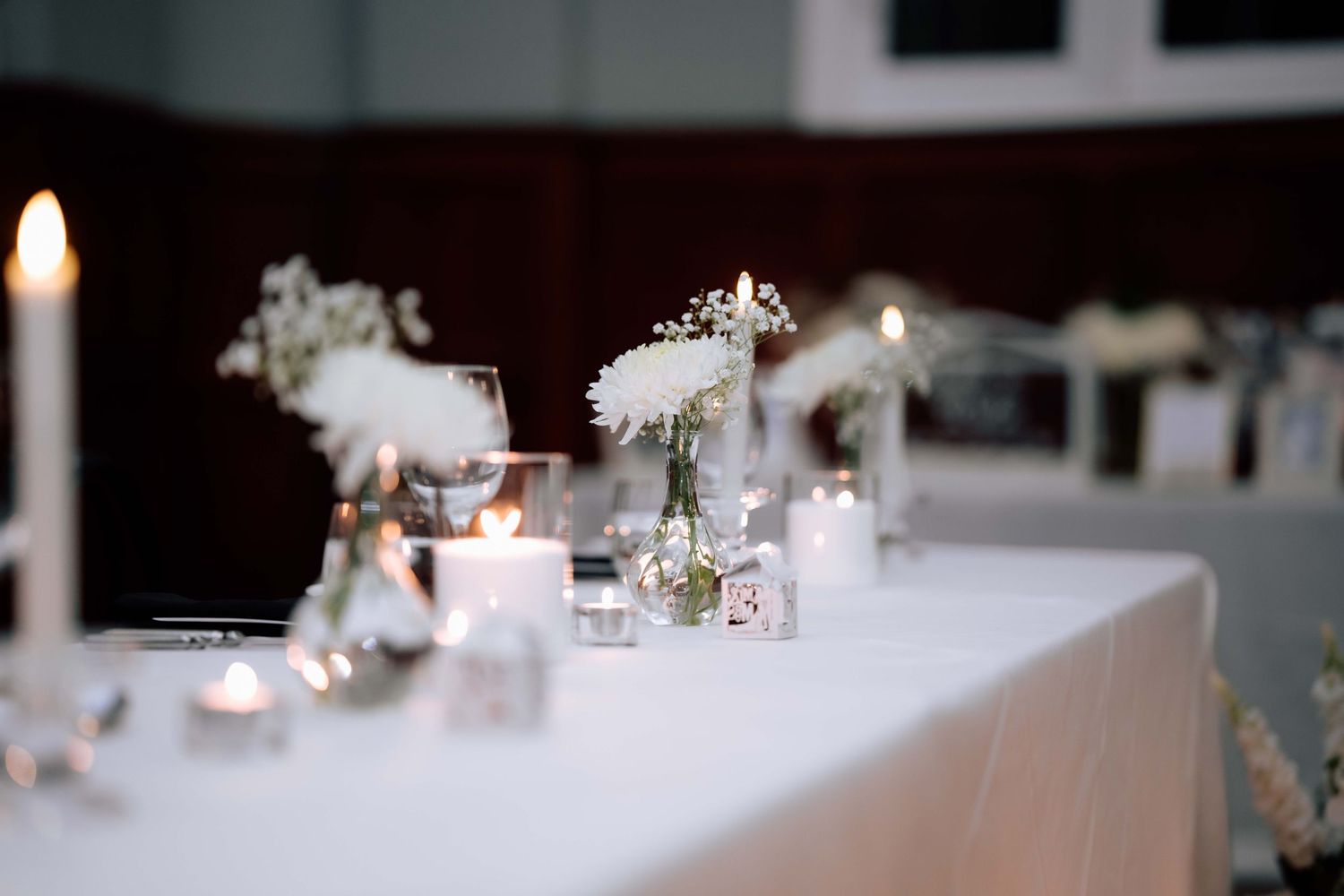 Elegant wedding reception table with white floral arrangements, candles, and glassware centerpieces.