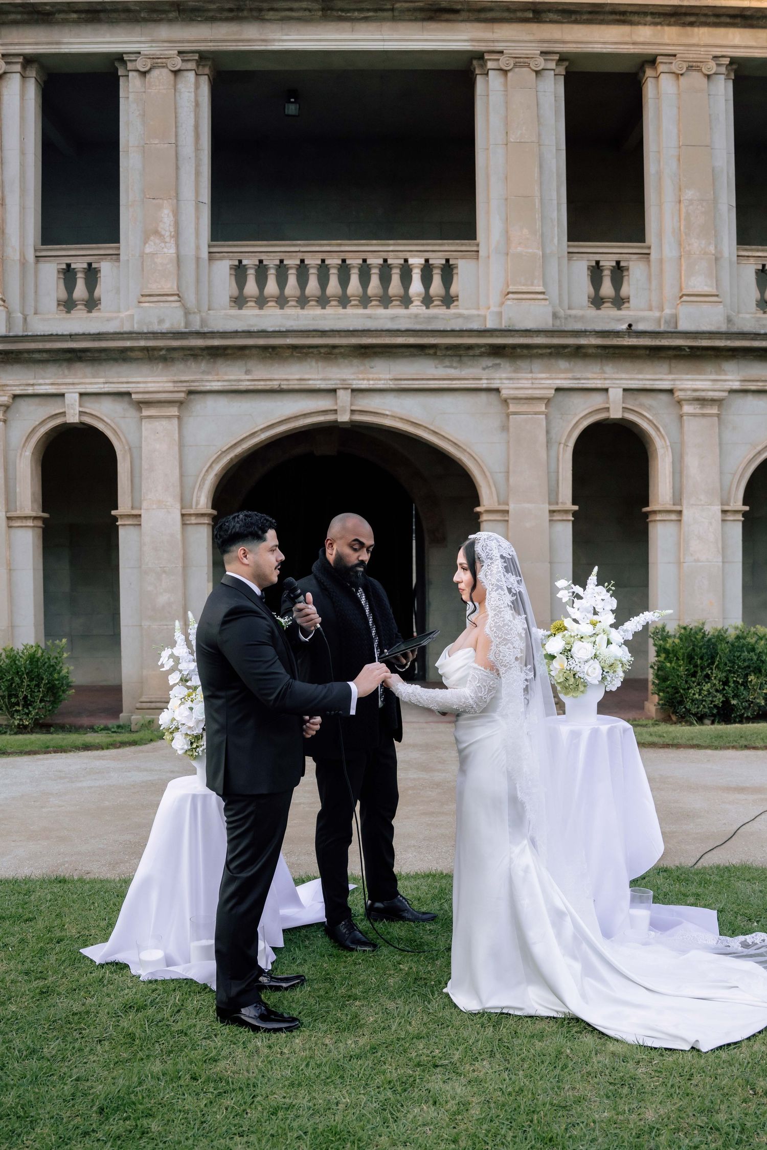 Bride and groom exchange vows outdoors before a grand classical stone building.