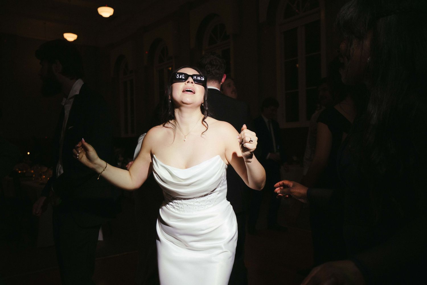 Bride wearing sunglasses dances freely in white gown at fun and energetic wedding reception.