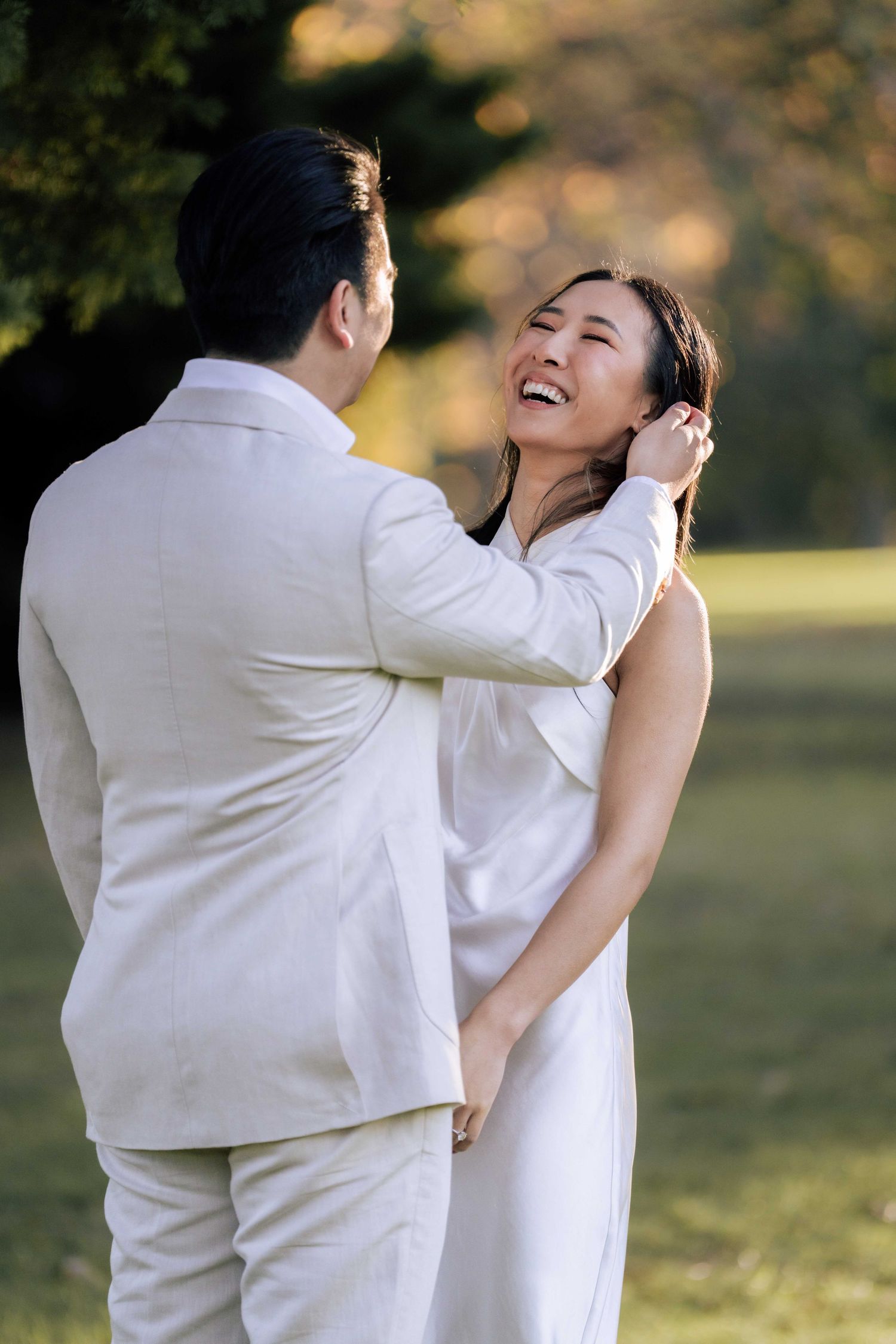 Joyful Asian couple in white outfits share a tender moment in a sunlit park, groom gently touching bride's face.