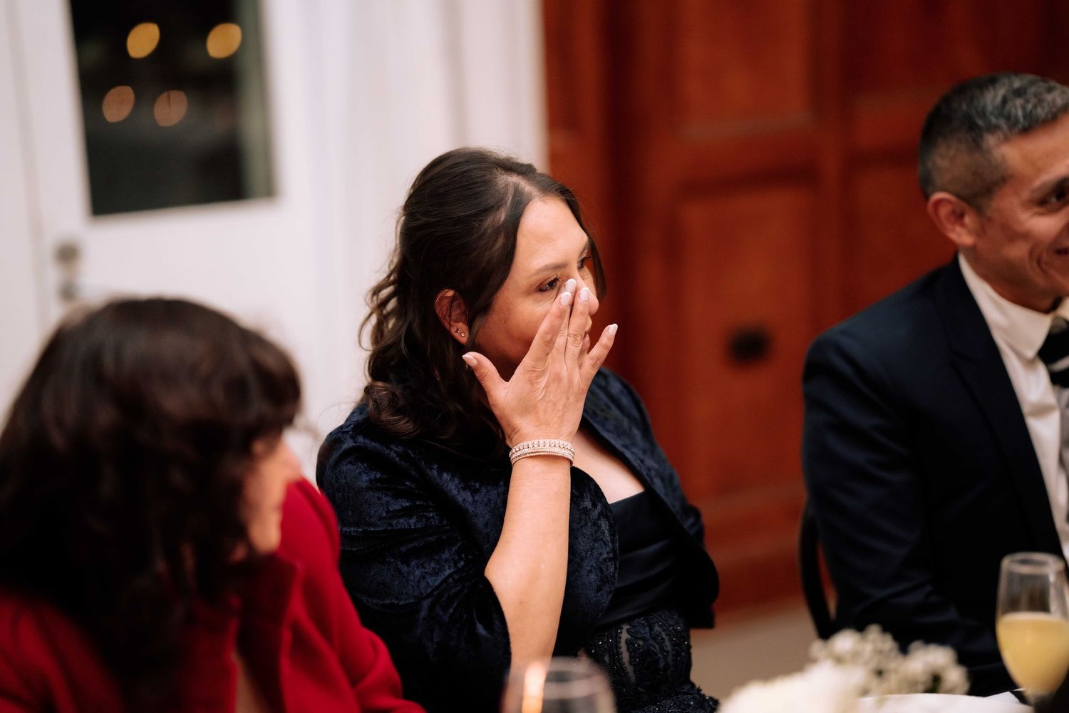 An emotional wedding guest covers her mouth in surprise while seated at a reception dinner table.