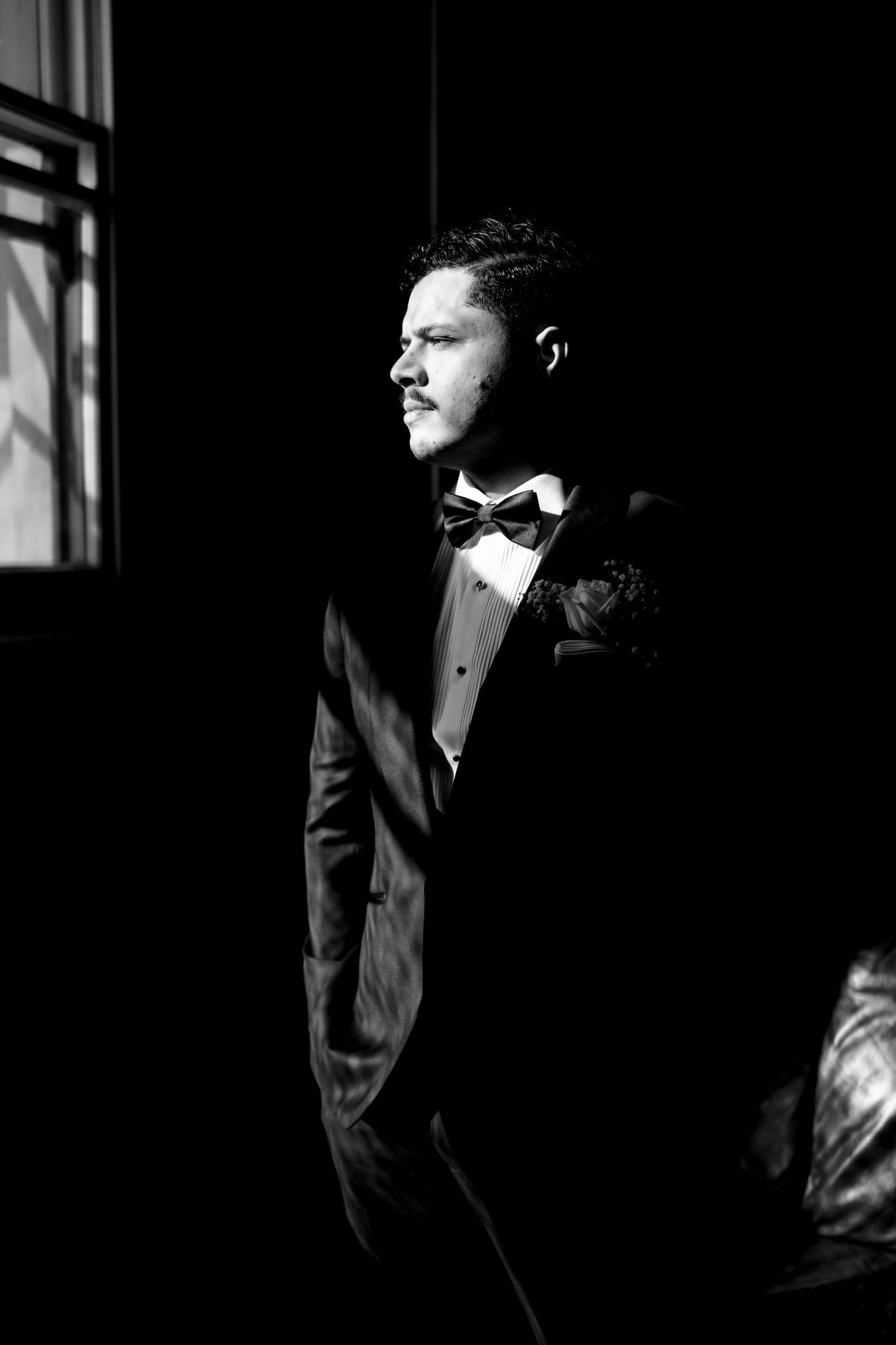 Groom in black tuxedo and bow tie standing by window in dramatic black and white wedding portrait.