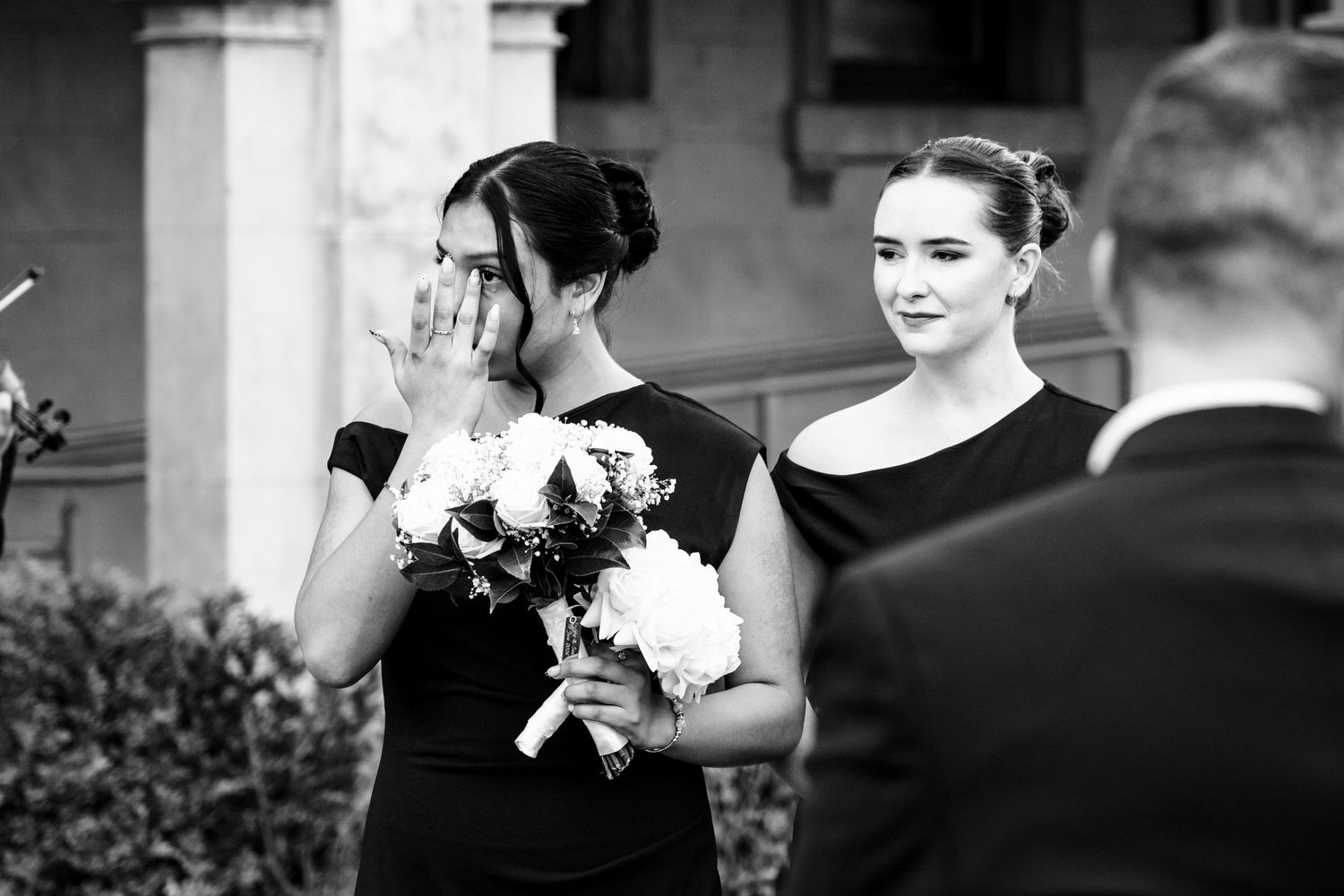 Emotional bridesmaid in black dress covering her face while holding white bouquet at wedding ceremony.