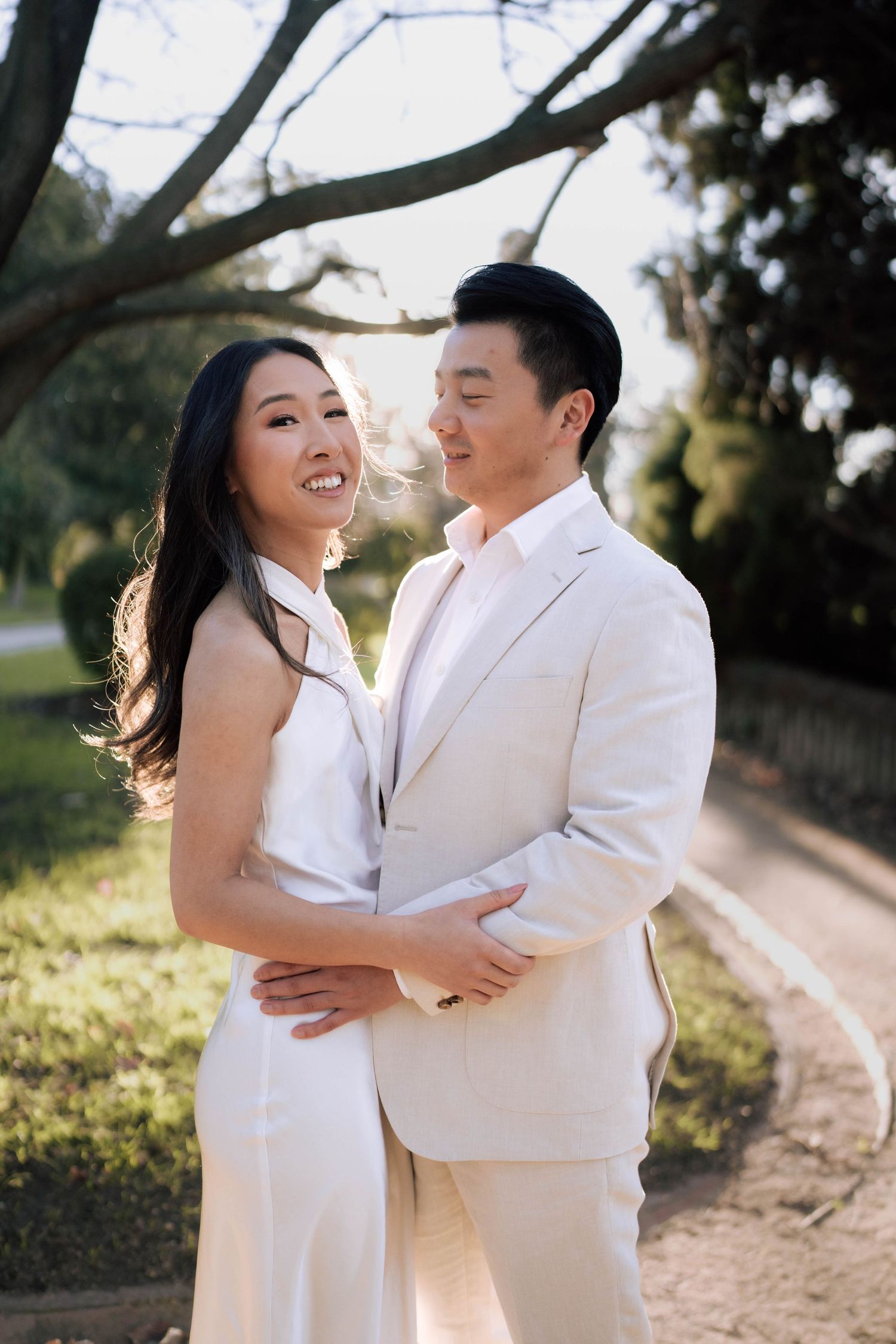 Couple in white outfits embracing outdoors, smiling, with sunlit trees and a garden path in the background.