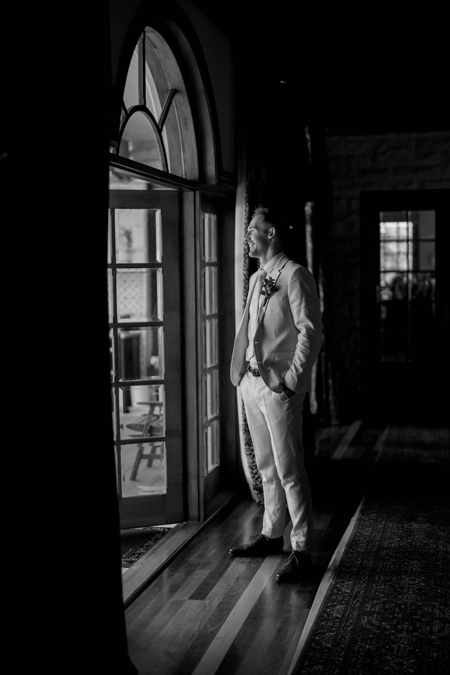 Black and white portrait of groom in white suit standing by arched window in venue.