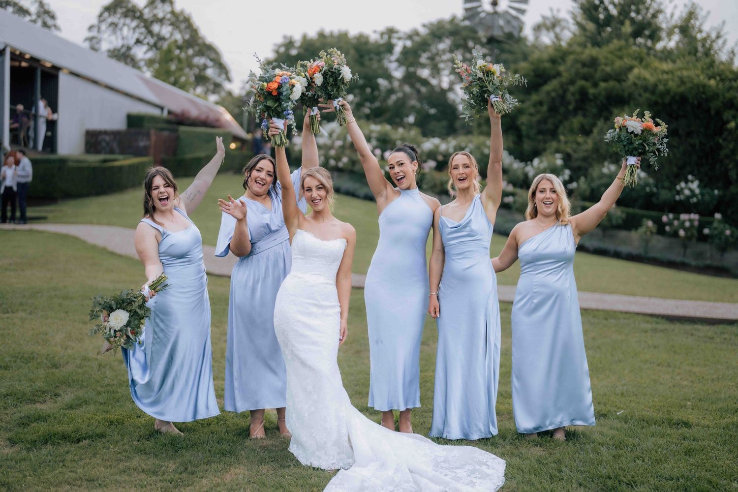 Bride in white gown celebrates with five bridesmaids in light blue dresses raising bouquets on garden lawn.