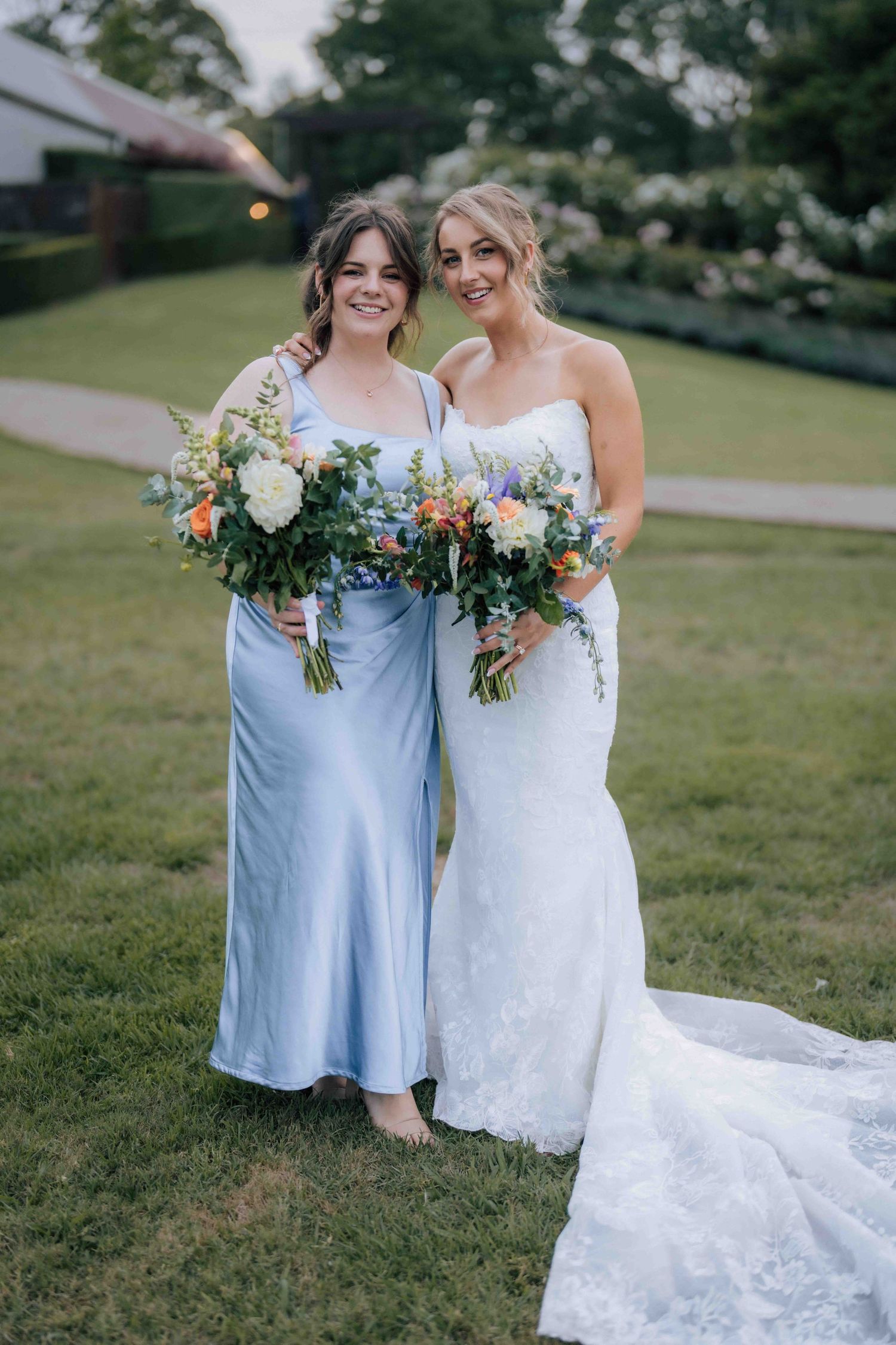 Smiling bridesmaid in light blue satin dress stands beside bride in white lace gown holding vibrant floral bouquets.