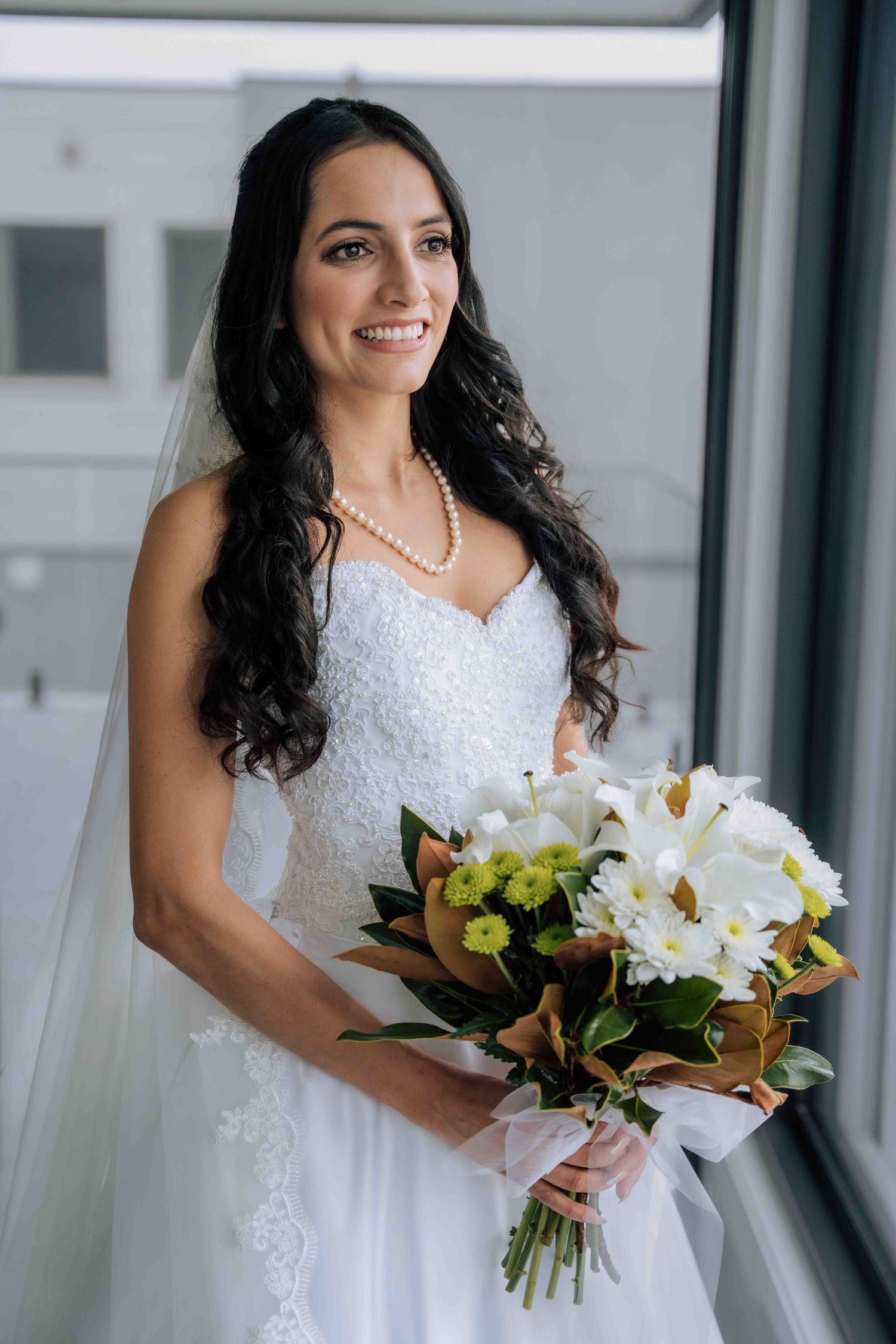 Smiling bride in white lace gown holding bouquet of white and yellow flowers by window.