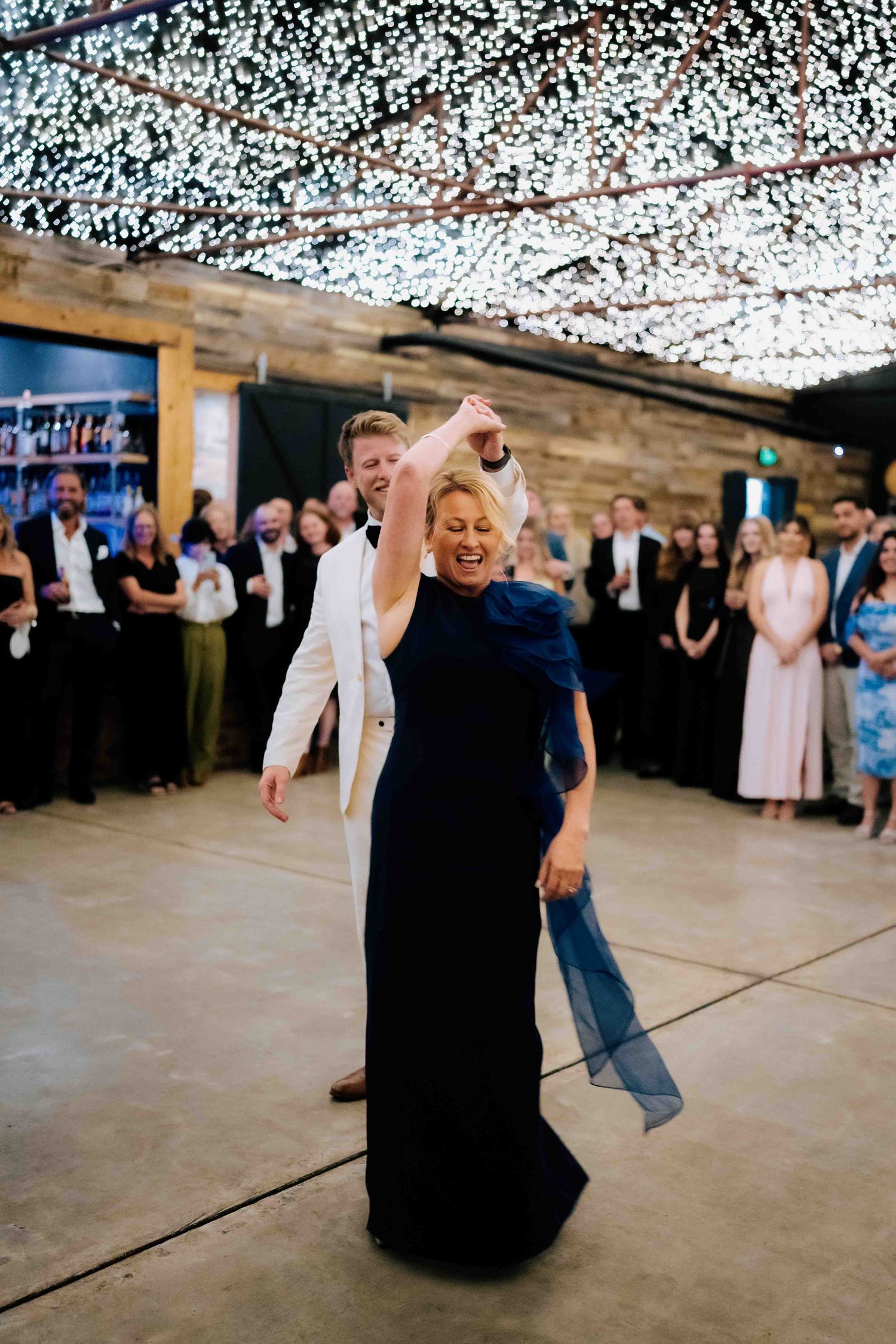 Woman in navy gown spins on dance floor under fairy lights at wedding reception.