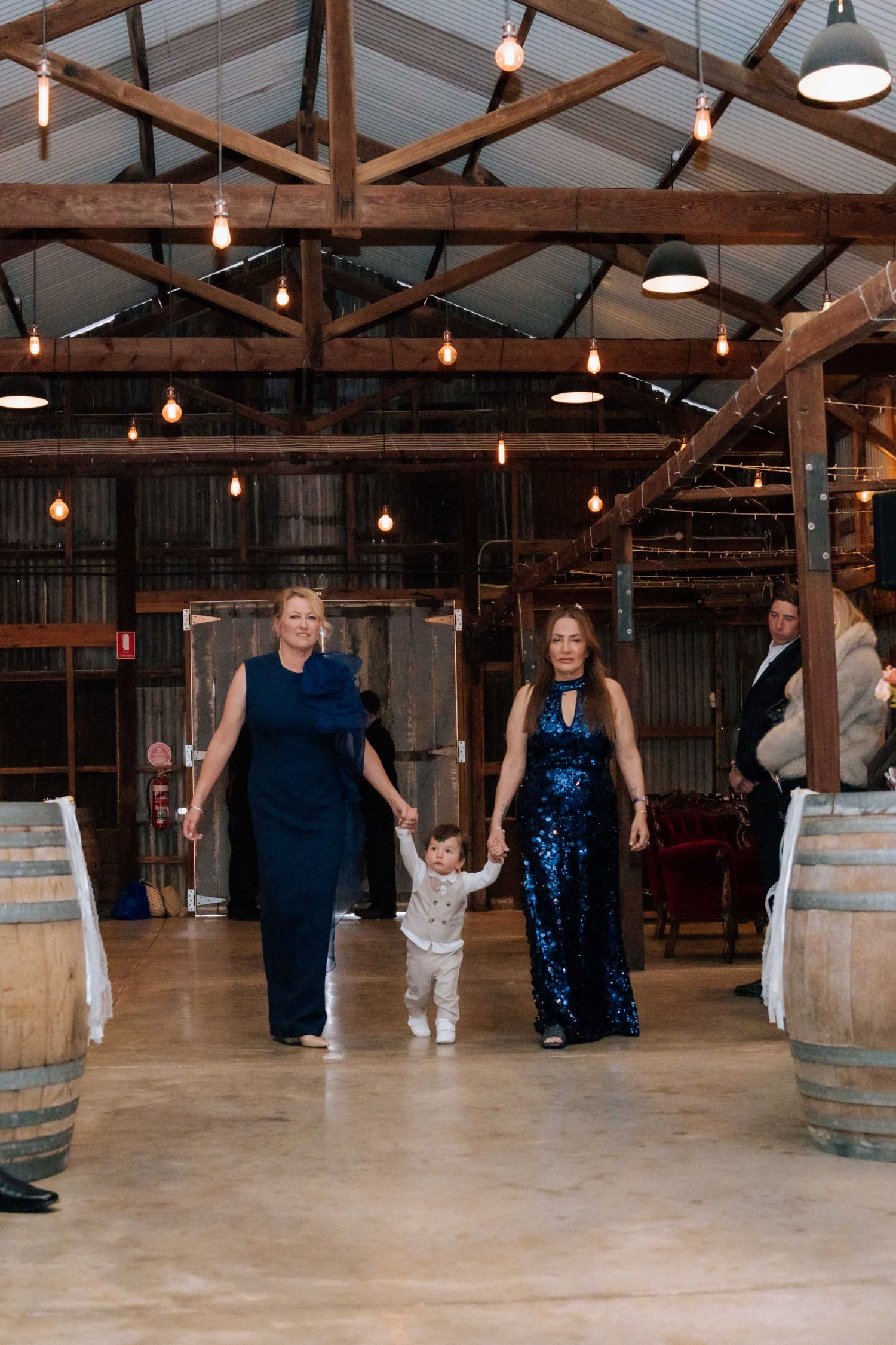Two women and a toddler walk hand-in-hand through a rustic barn wedding venue with warm Edison lights.