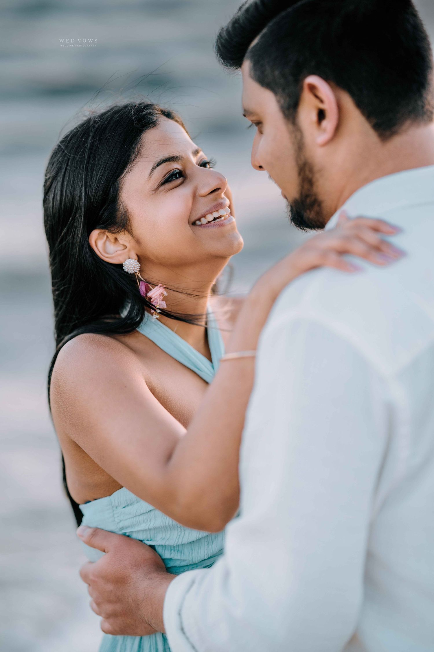 Smiling Indian woman in mint dress embracing man in white shirt at beach during sunset.