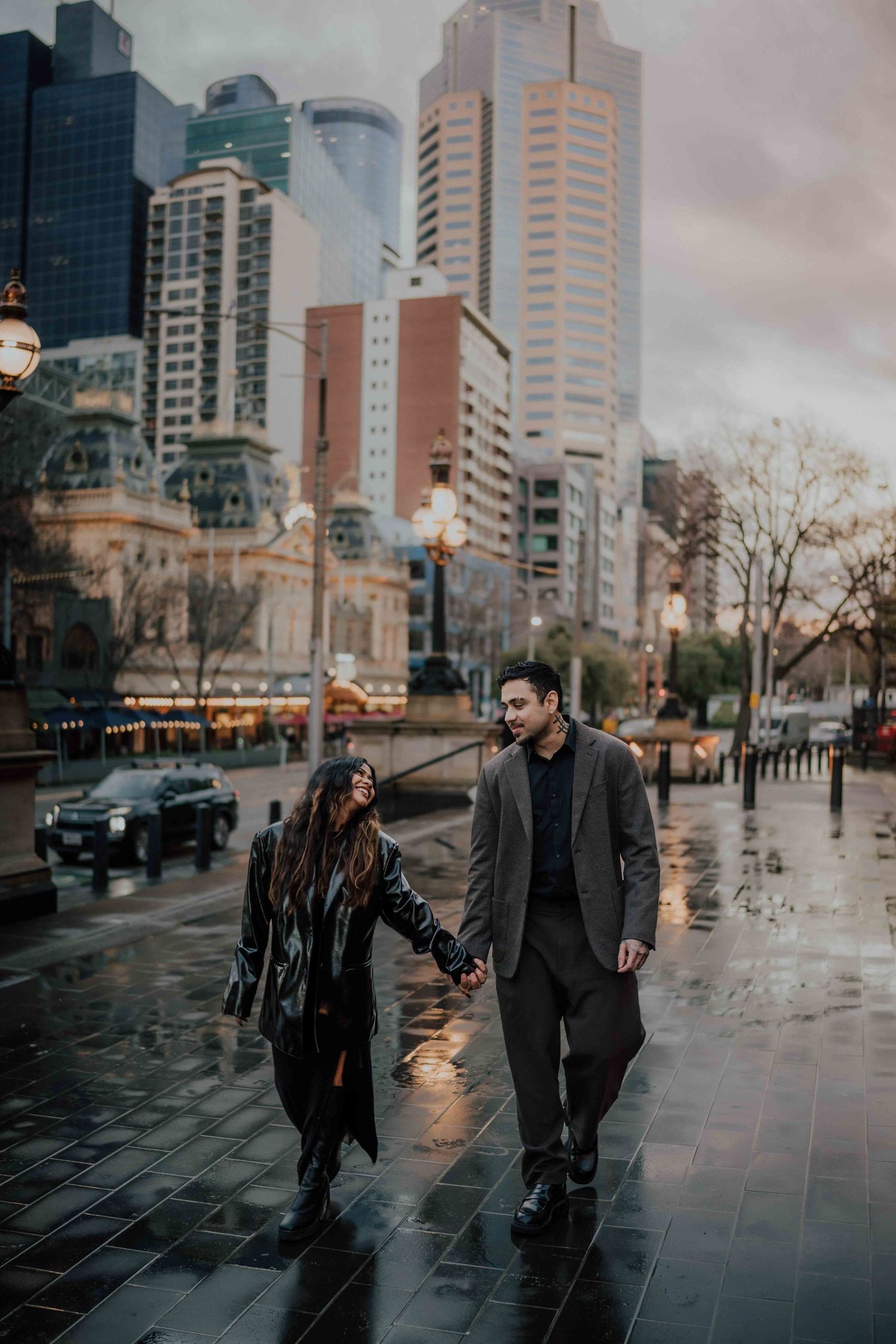 Couple holding hands walking on rain-slicked city streets with Melbourne skyscrapers in background.