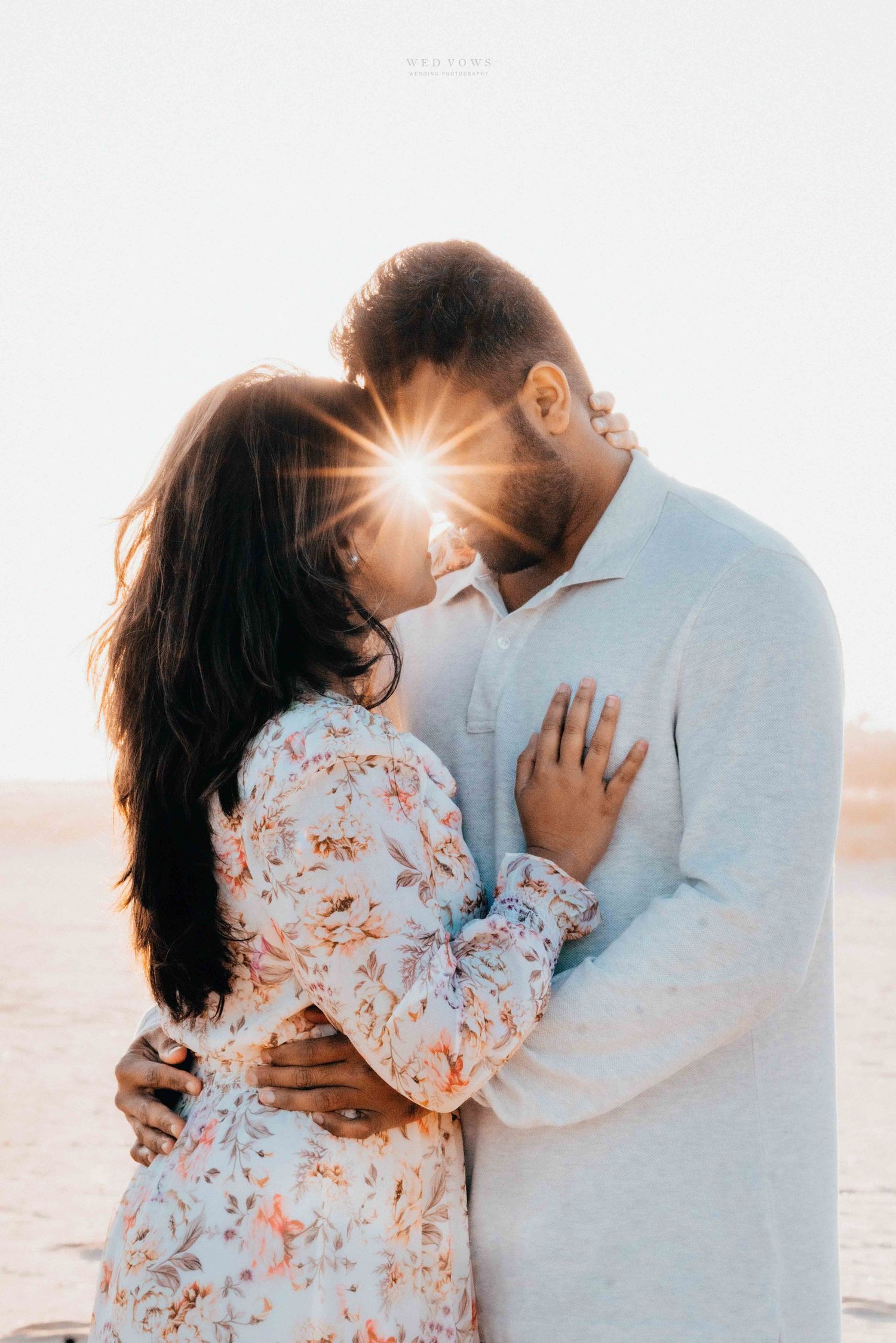 Couple embracing with a sunburst between them during a golden hour beach photoshoot.