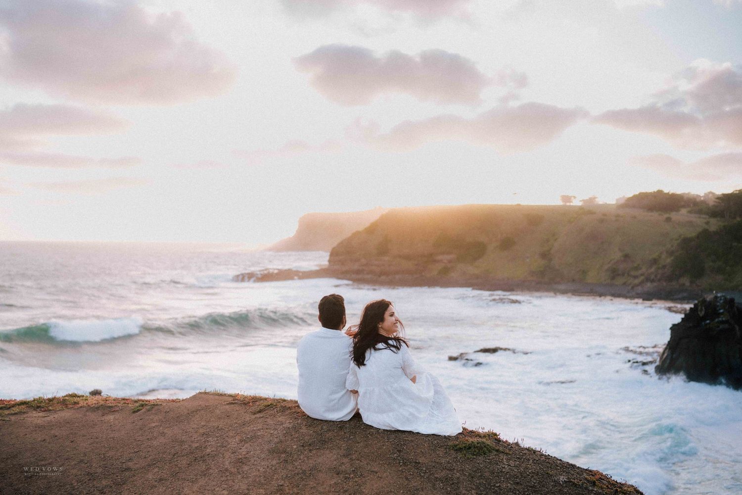 Couple in white sitting together on cliff overlooking ocean waves at colorful sunset.