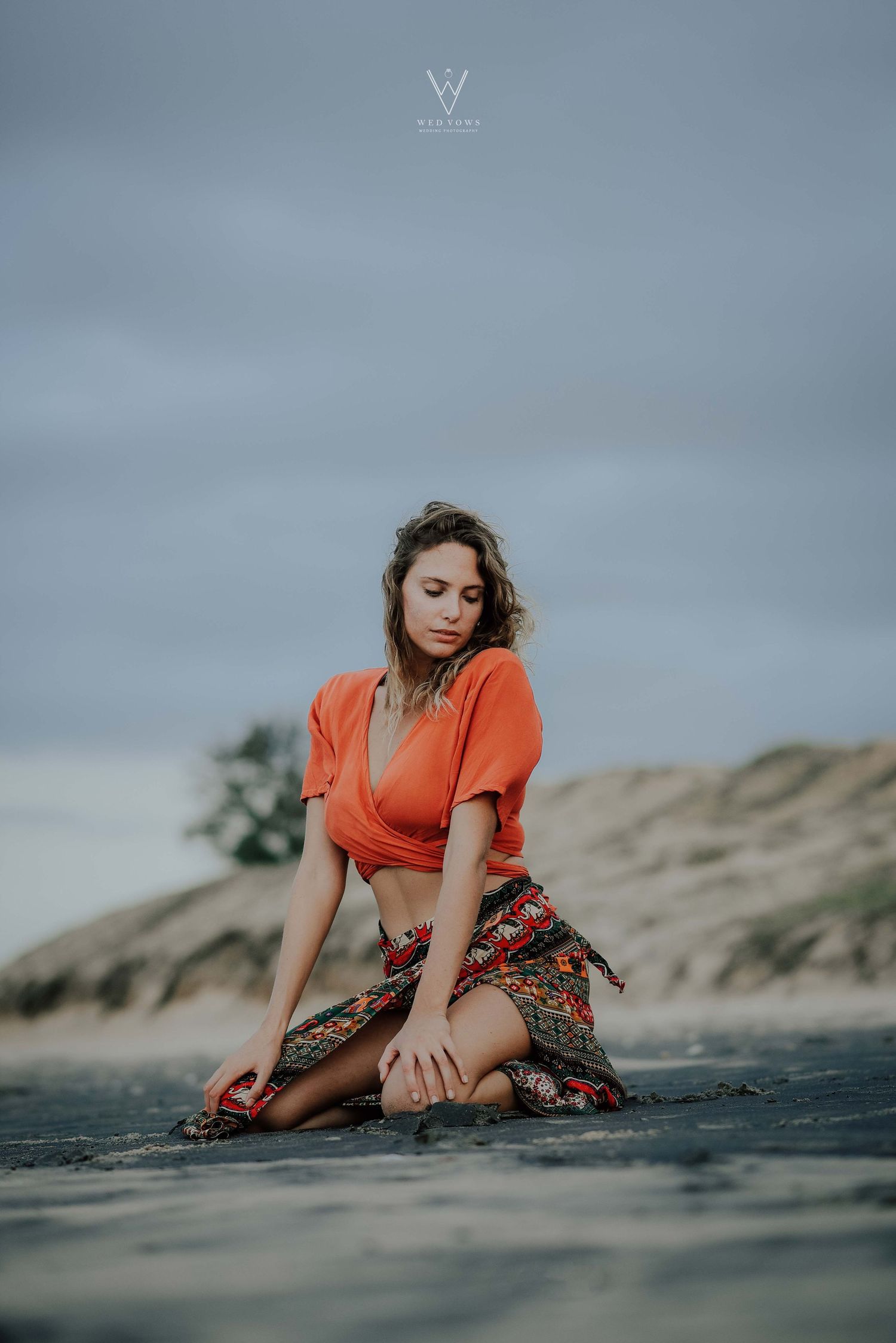 Woman in orange crop top and patterned skirt posing gracefully on a sandy beach.