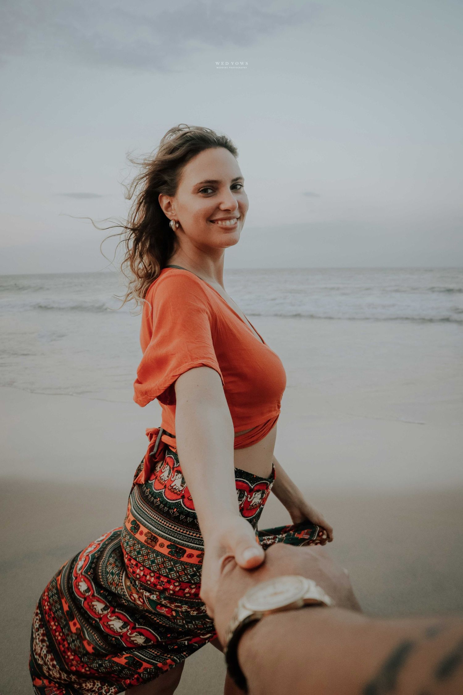 A smiling woman in an orange crop top and boho skirt holds hands with someone on a breezy beach.