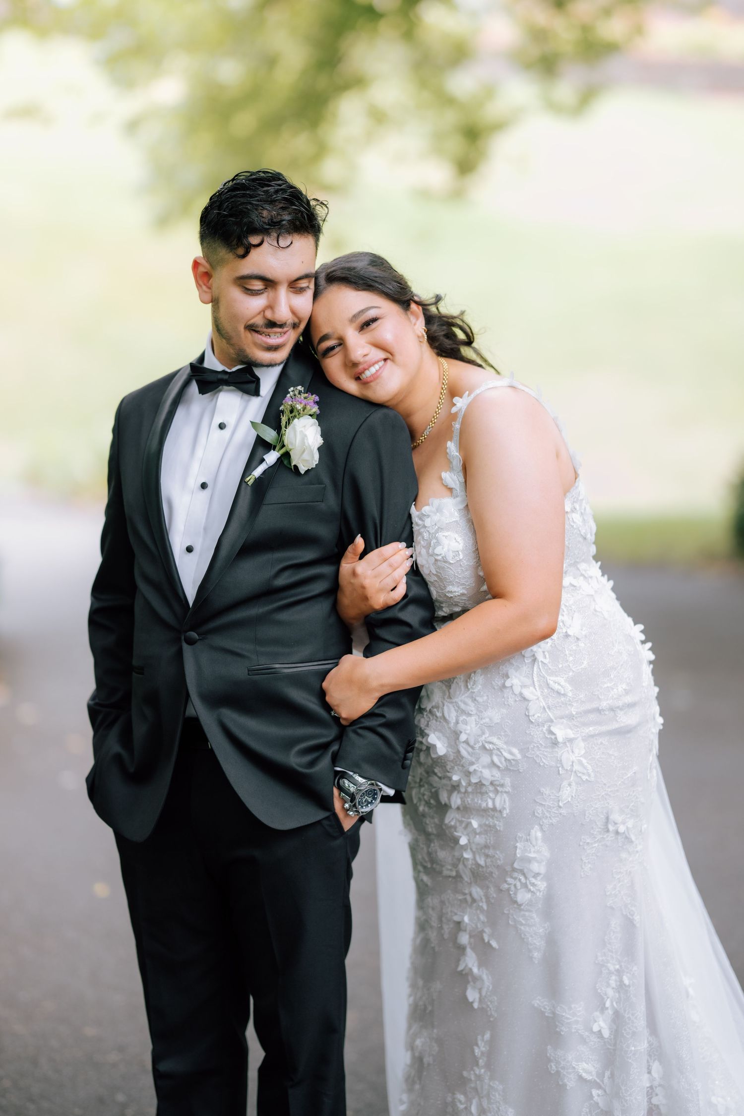 Bride in lace gown embraces groom in black tuxedo, smiling joyfully during outdoor wedding portrait.