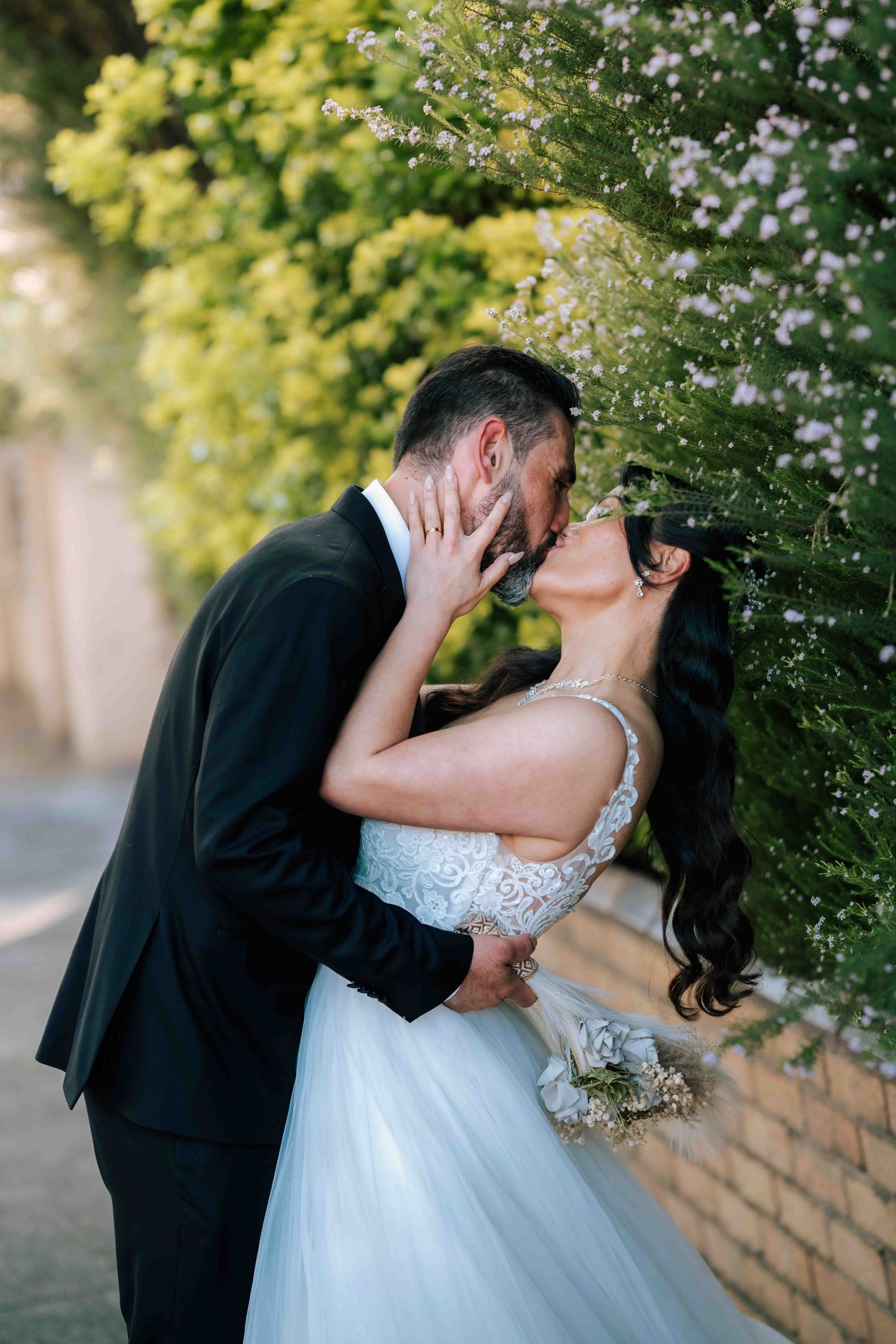 Bride and groom sharing romantic kiss beside blooming flower wall outdoors.