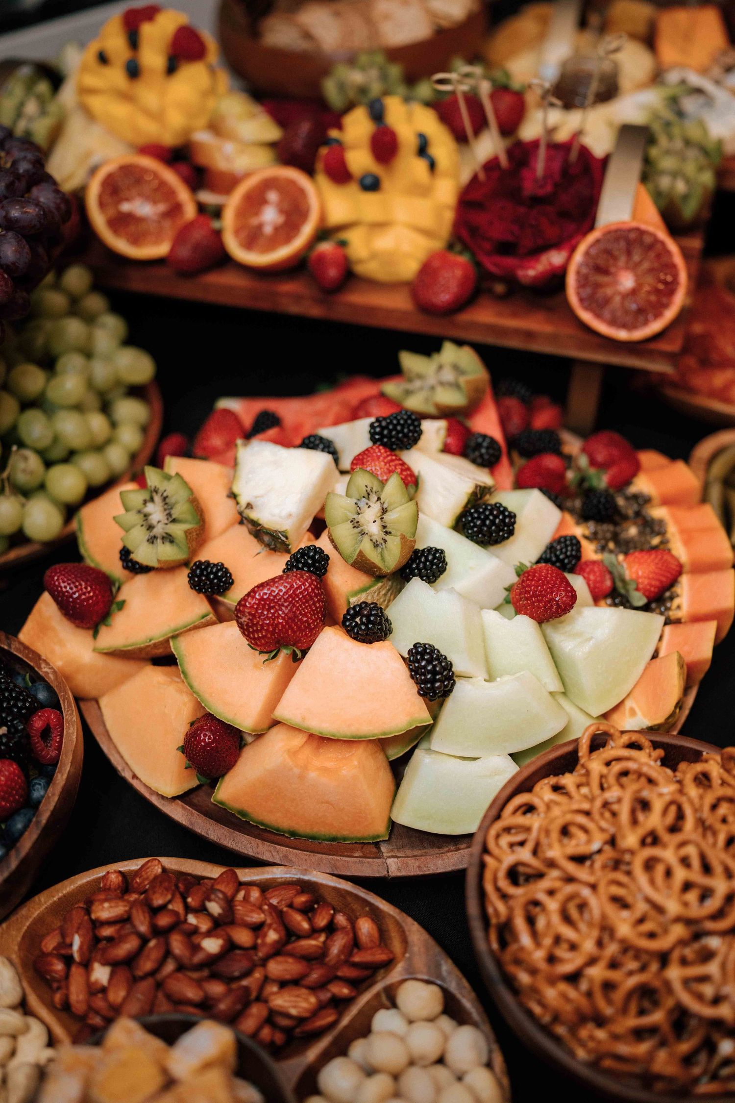 Elegant grazing table display with sliced melon, mixed berries, tropical fruits, and assorted nuts.