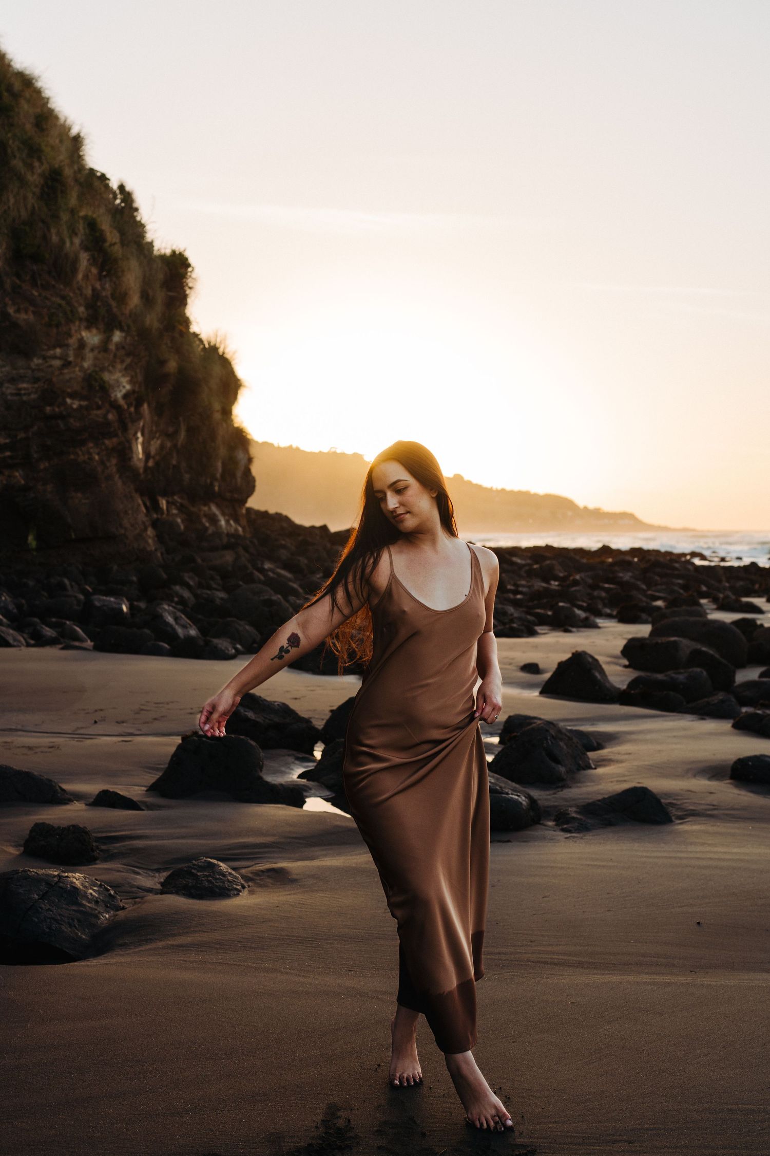 A woman holds her dress and dances across the black sand of a West Coast New Zealand beach. Image created by Photography by Chloe.