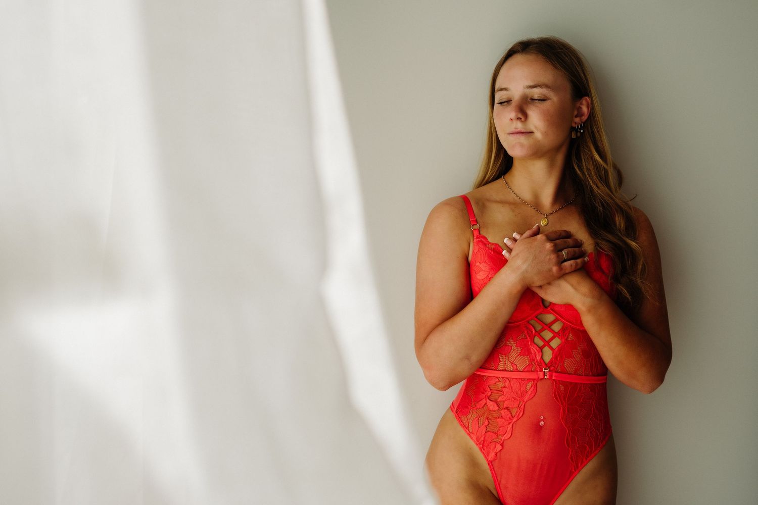 A woman holds her hands to her heart space, standing in soft natural window light
