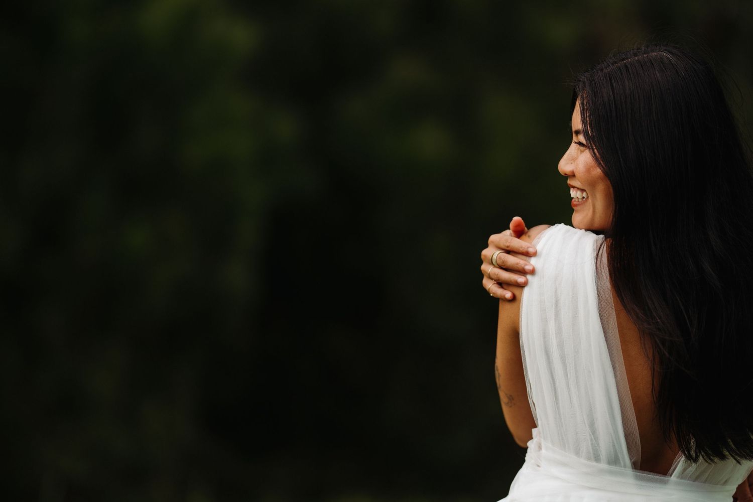Woman in a white dress smiles as she looks over her shoulder, while standing in an orchard in Raglan New Zealand, by portrait photographer