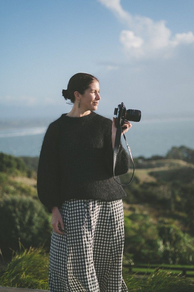 Woman holds a camera, smiling while looking at the ocean, into sunlight