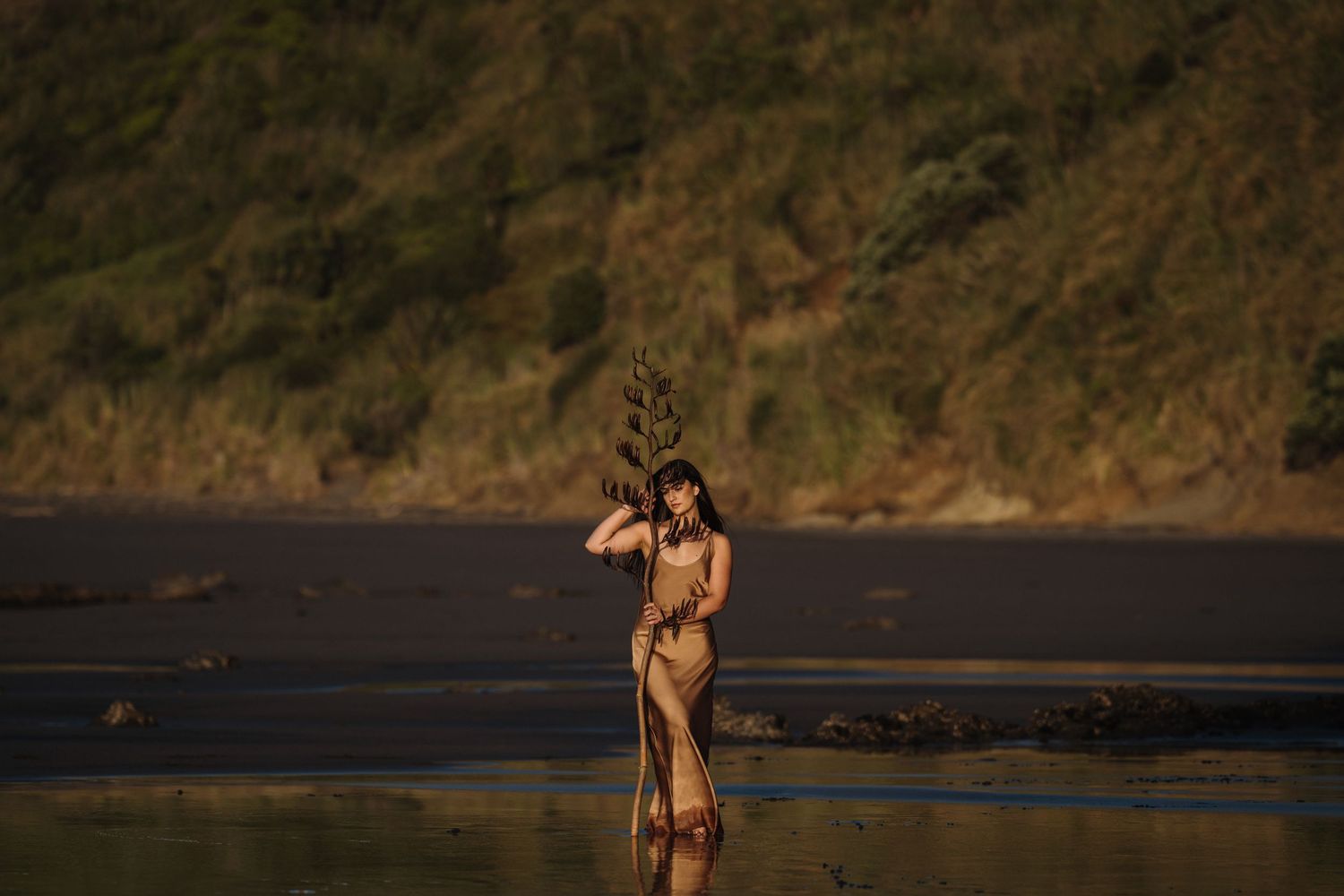 A woman holds a flax branch standing in the sunset on a West Coast, black sand beach. Image created by Photography by Chloe
