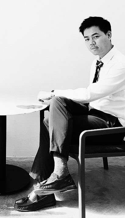 Black and white portrait of figure in business attire sitting in chair against white wall, wearing dress shirt and tie.