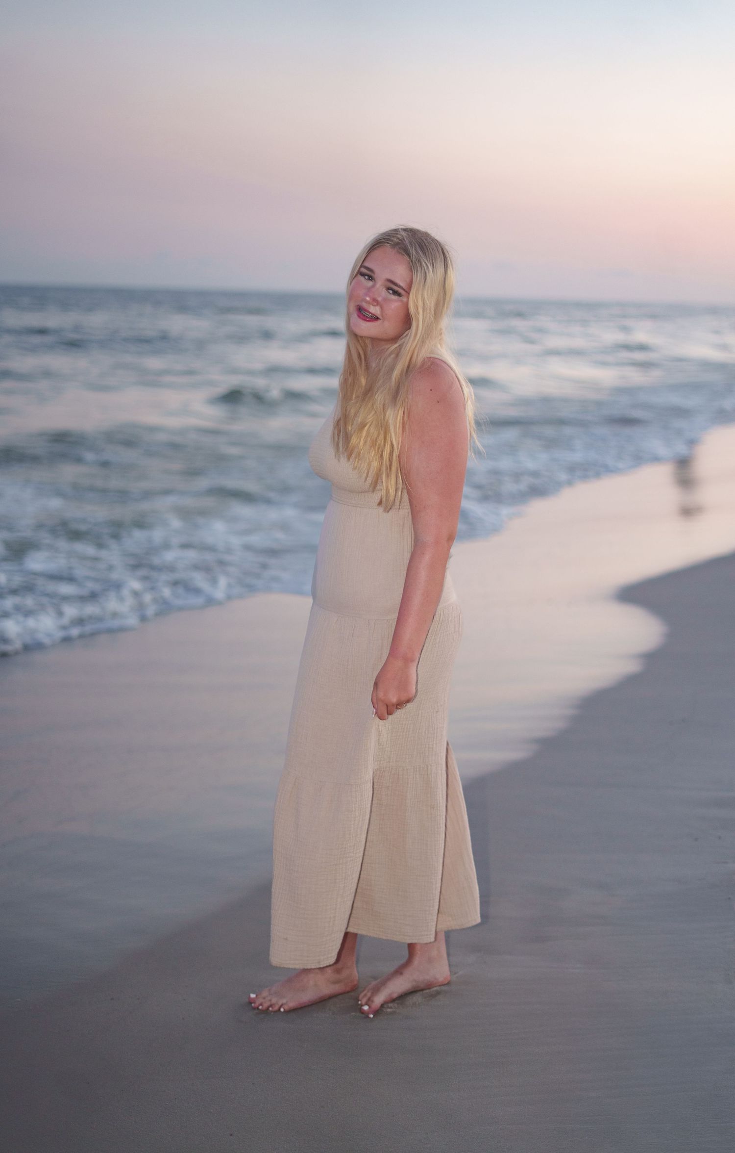 Blonde woman in a beige maxi dress standing barefoot on a sandy beach at sunset.
