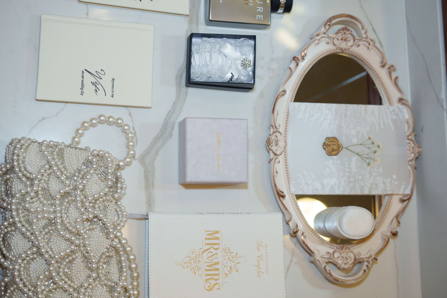 Elegant bridal accessories display featuring pearl clutch, ornate mirror, jewelry boxes, and stationery on marble surface.