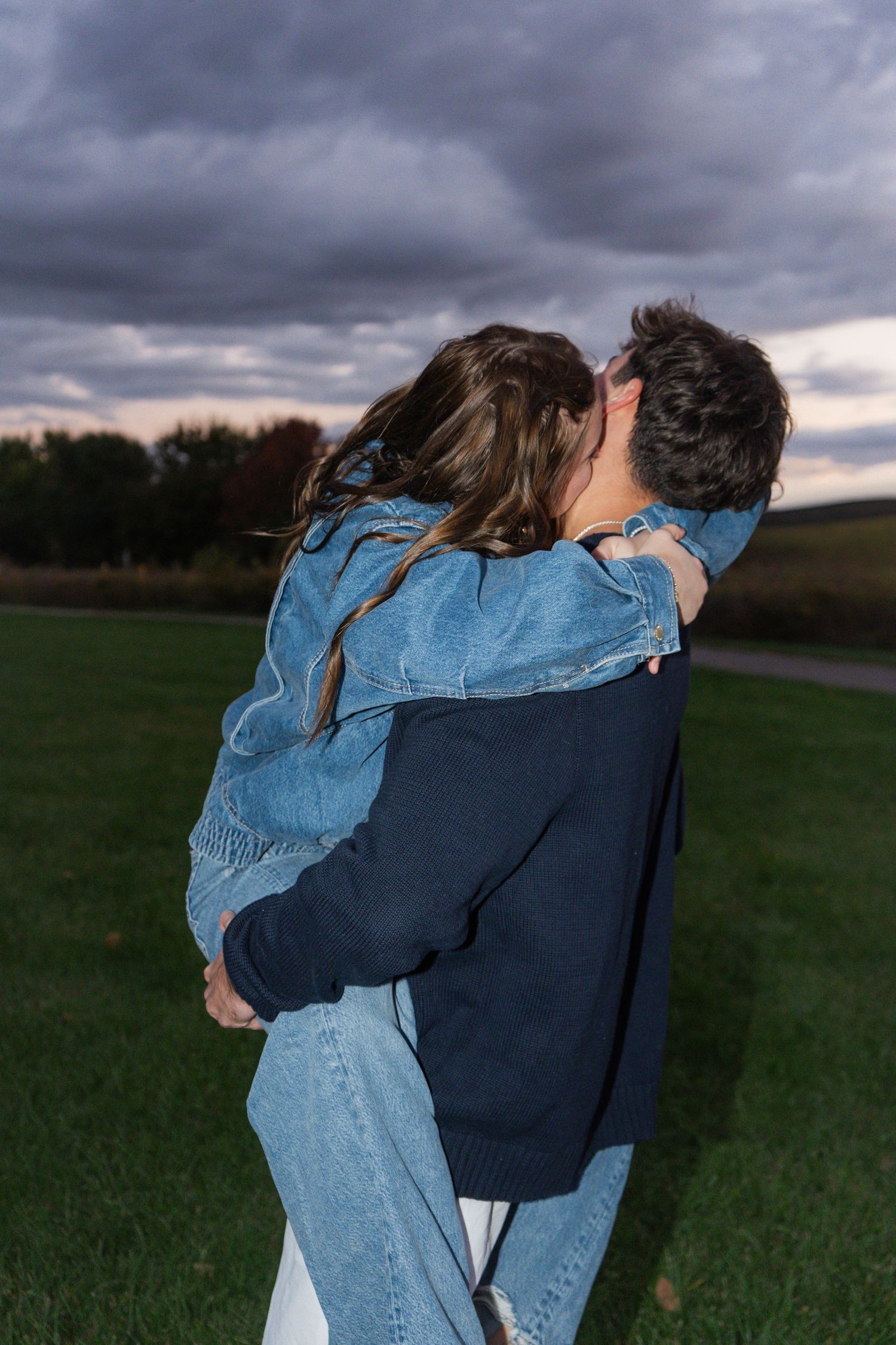 A couple embraces in a field at dusk with dramatic cloudy skies overhead.