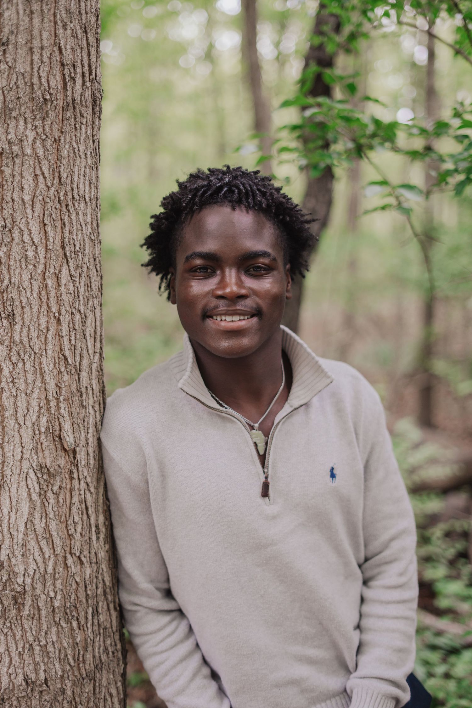 Young man in beige Ralph Lauren zip pullover smiling while leaning against a tree in a lush green forest.
