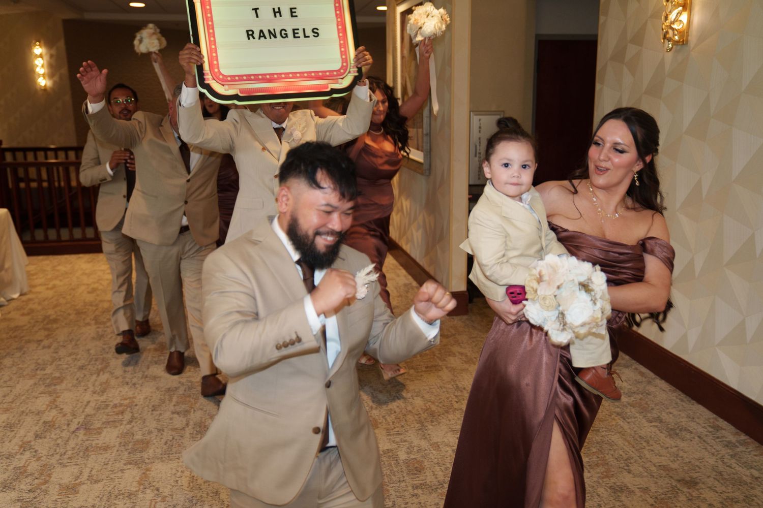 Wedding party celebrating with groom dancing, bride holding child, guests cheering under a personalized sign reading The Rangels.