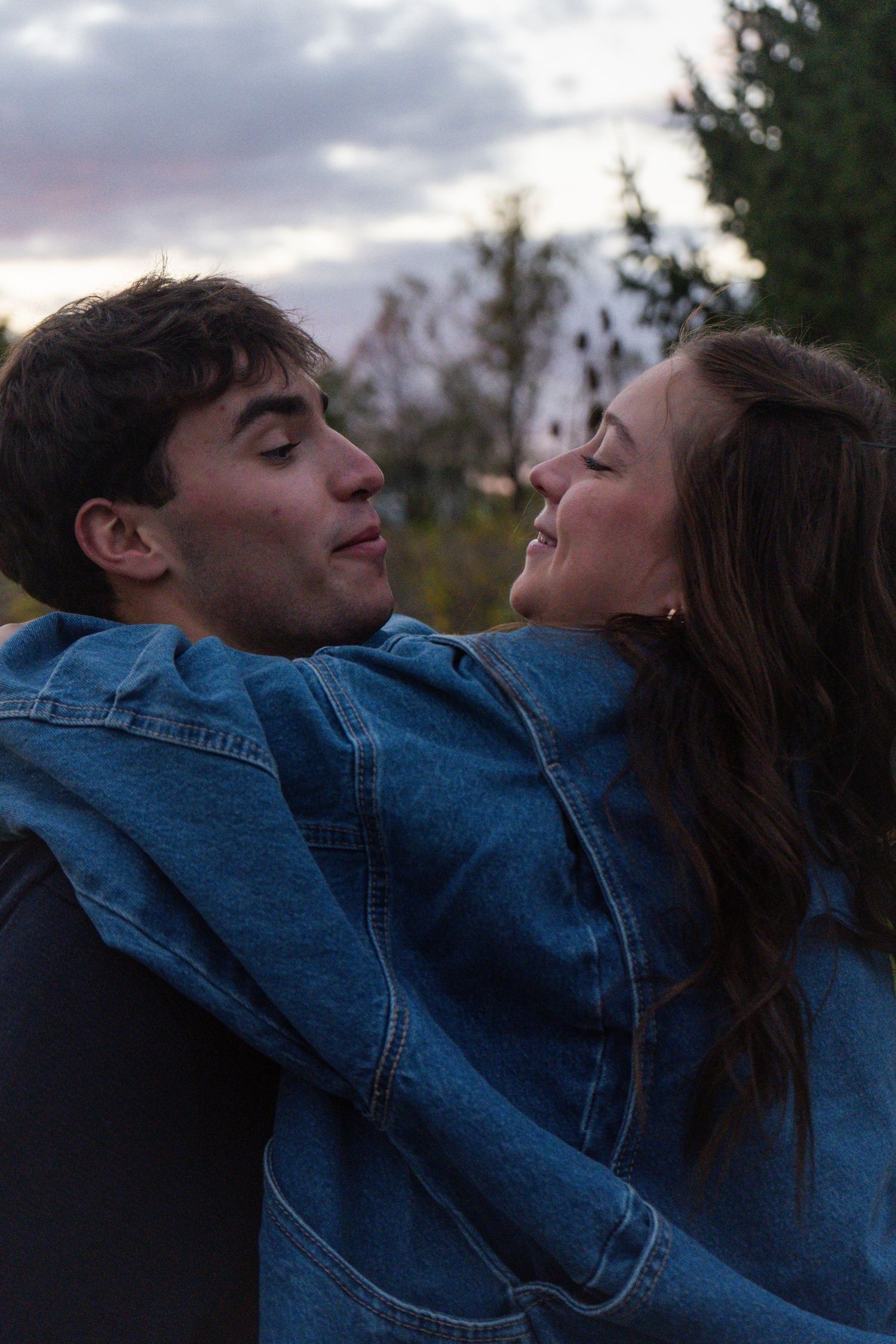 Two people in denim jackets lean back together against a cloudy sky in a romantic moment.