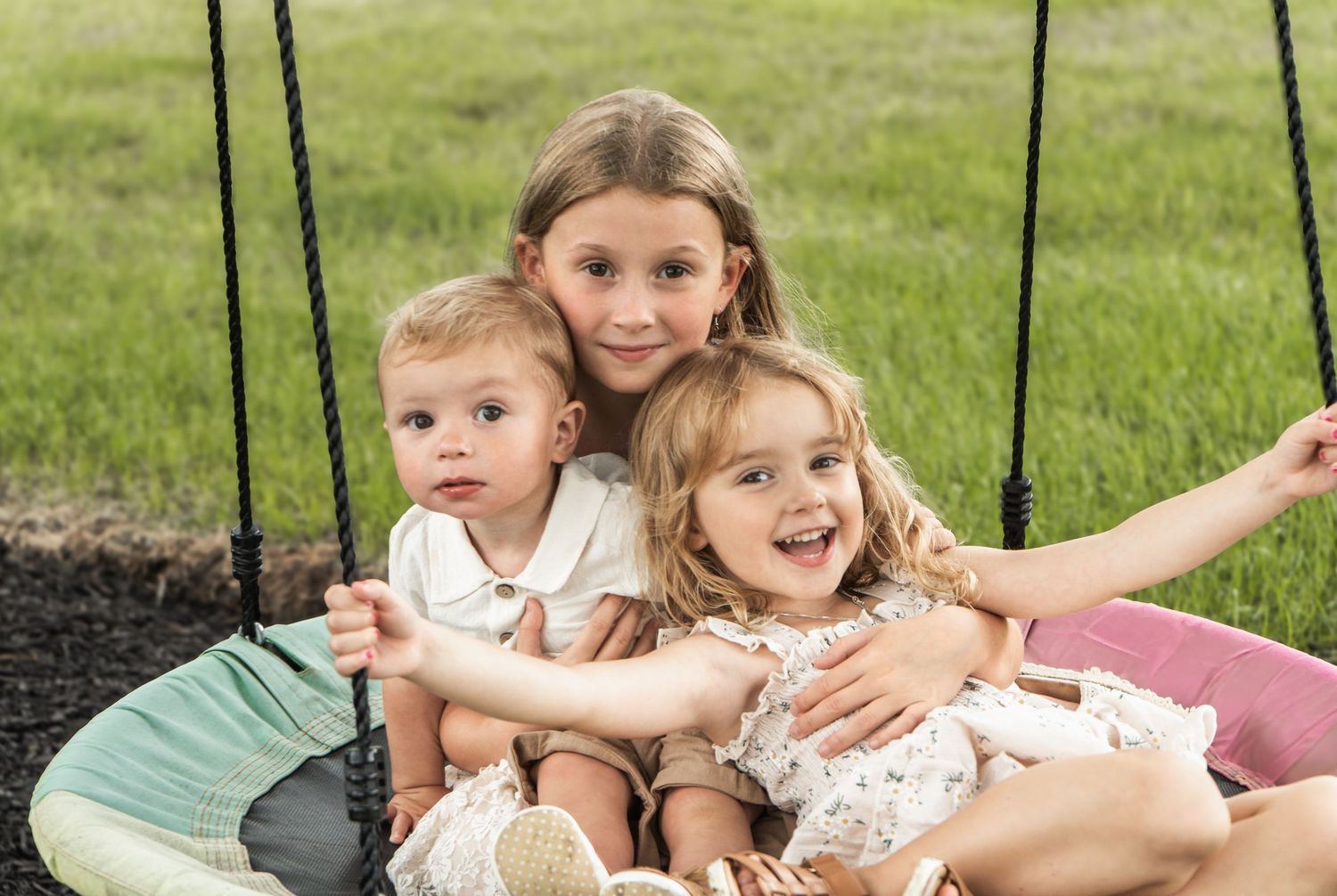 Three children sit close together on an outdoor swing with a grassy backdrop, laughing and enjoying themselves.