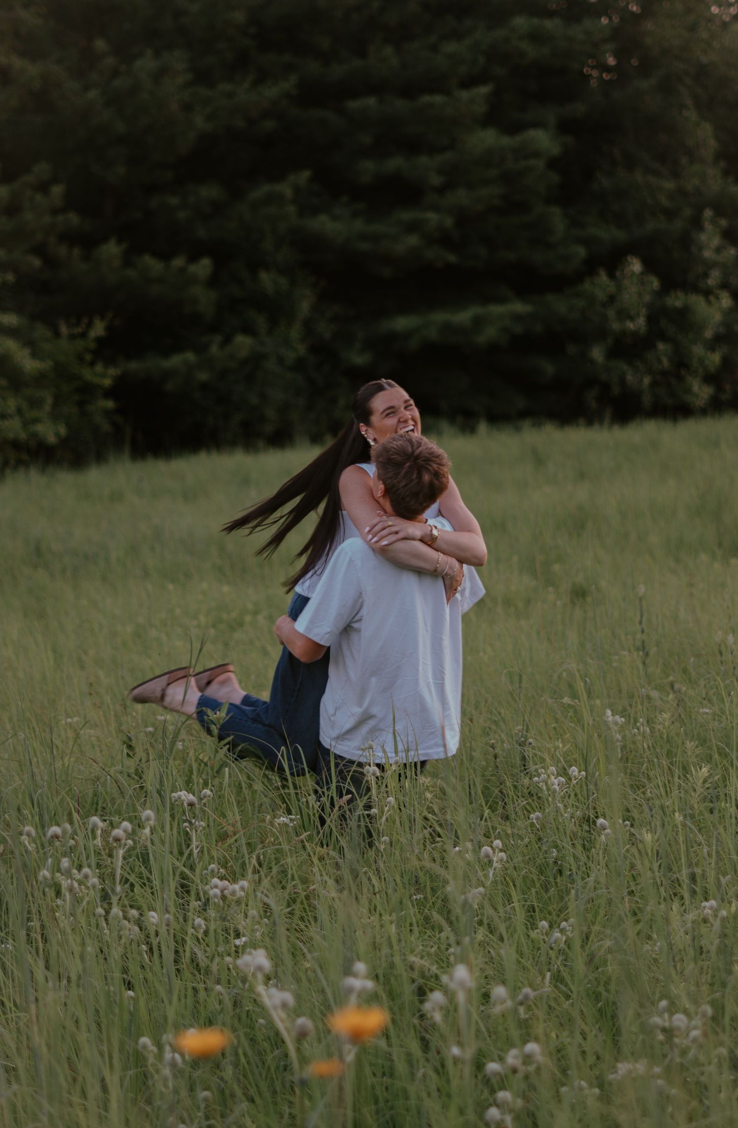 Couple embracing in a lush green meadow with wildflowers, sharing a romantic moment surrounded by trees.