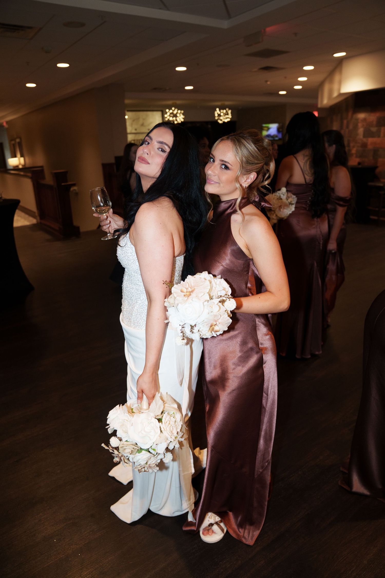 Bride in white gown and bridesmaid in mauve satin dress pose together holding bouquet at elegant wedding reception.
