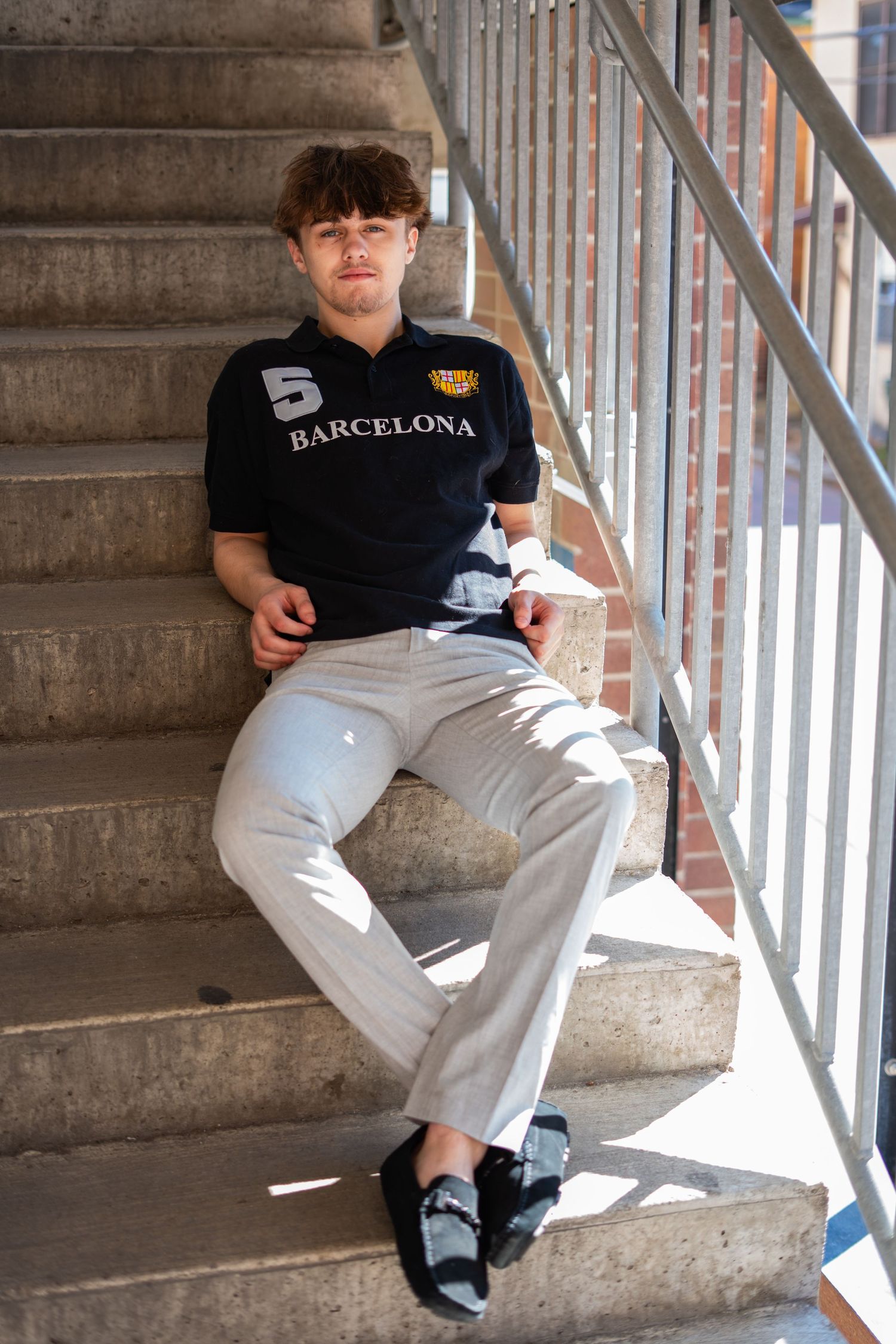 Young man in Barcelona polo shirt and white pants sitting on concrete stairs in sunlight.