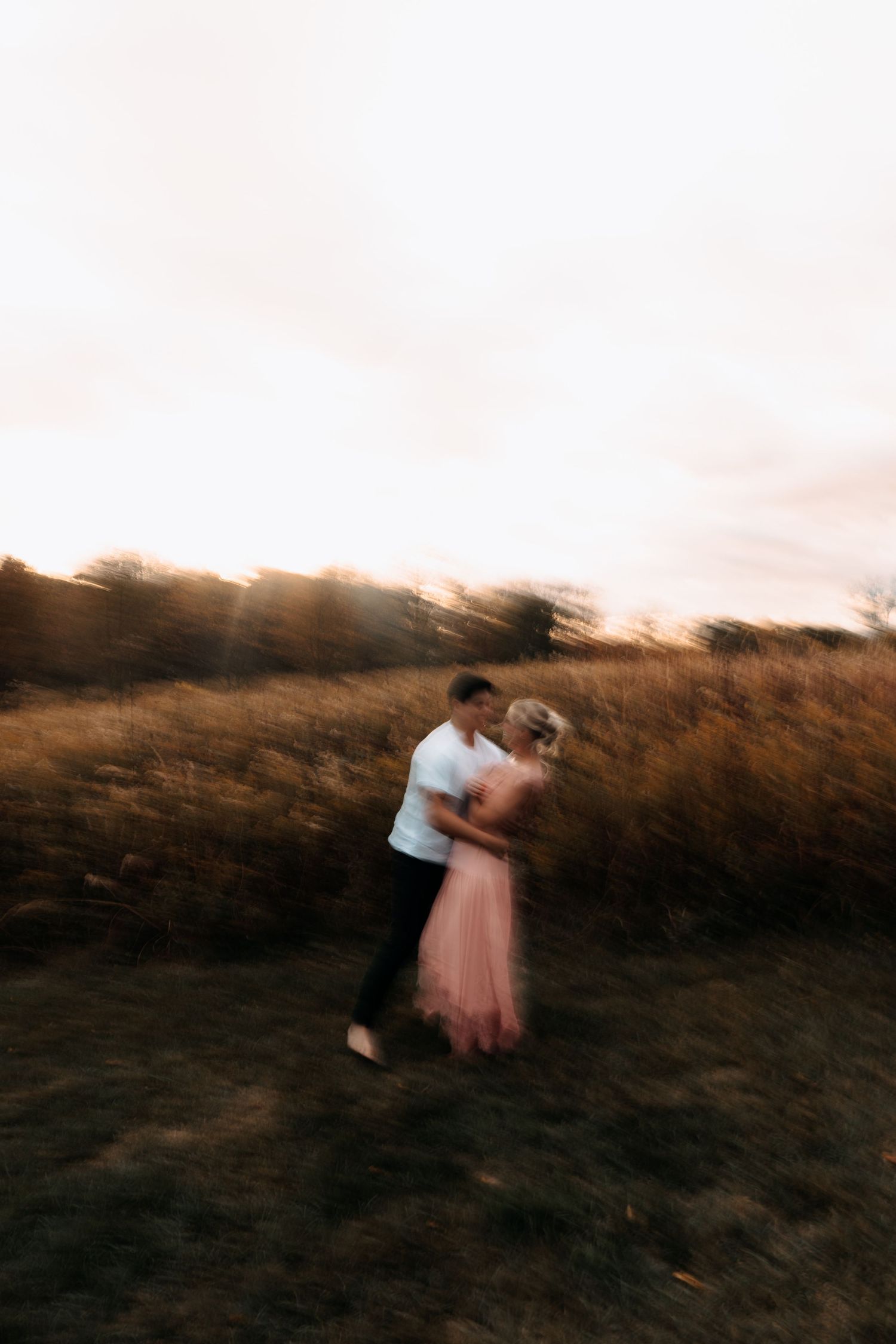 Couple embracing in a golden field at sunset, woman in pink dress, motion blur effect creating a dreamy romantic atmosphere.