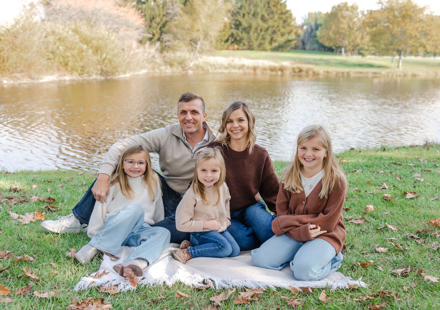 Family of five smiling together on a blanket by a serene pond during autumn, surrounded by colorful fallen leaves.