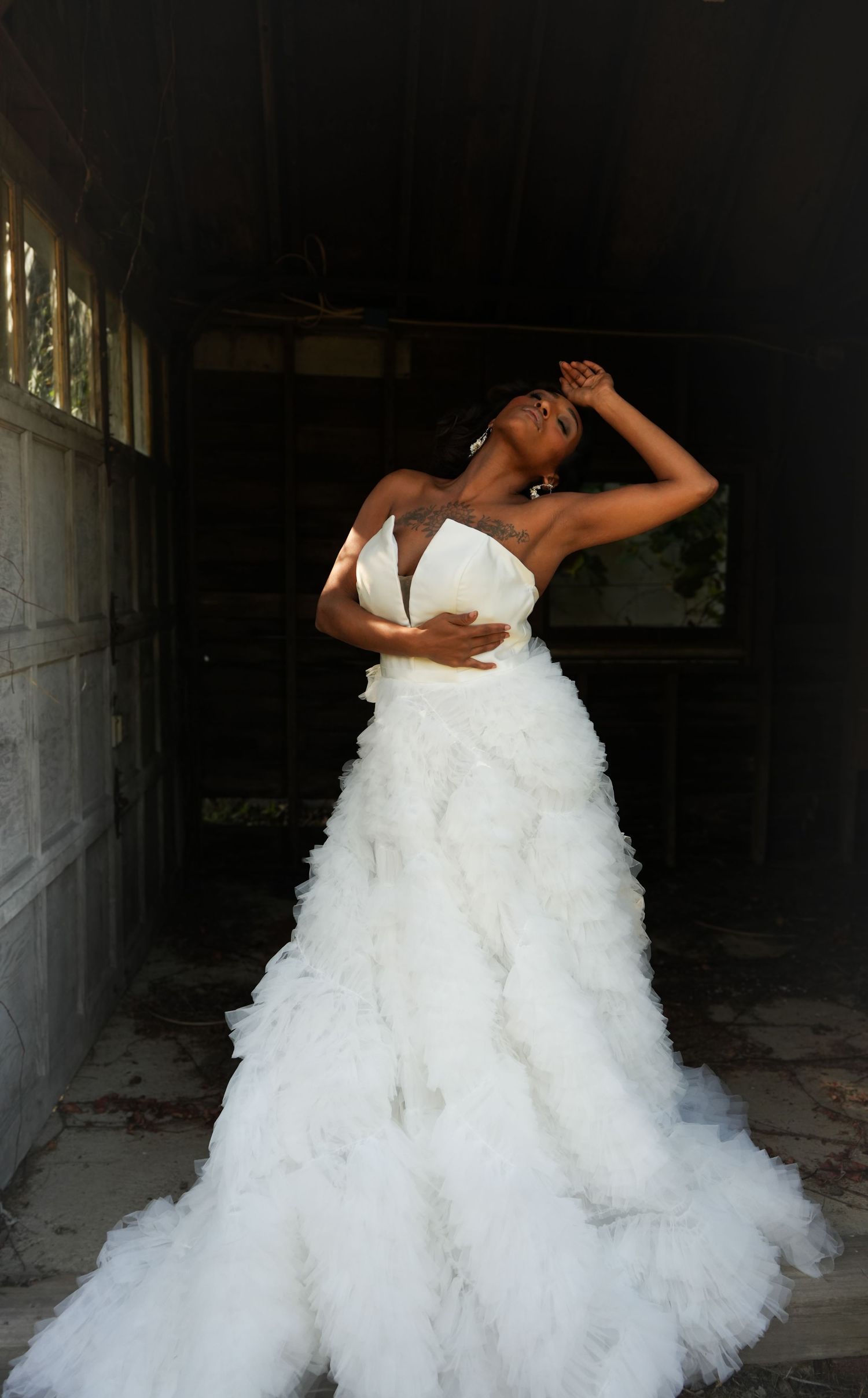 A dramatic bridal photoshoot in an industrial setting shows a flowing white wedding gown with ruffled tulle layers.