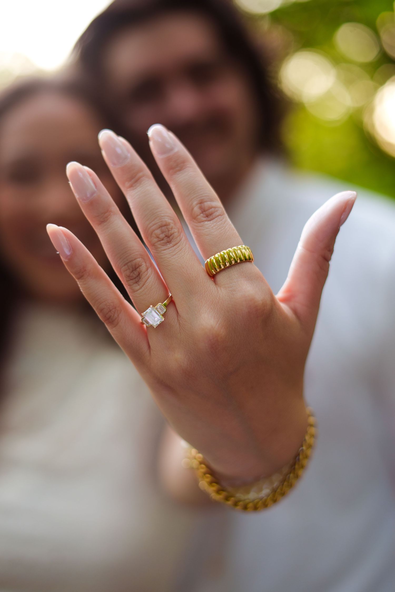 Close-up of a woman's hand displaying a diamond engagement ring and gold band, with a couple smiling in the background.