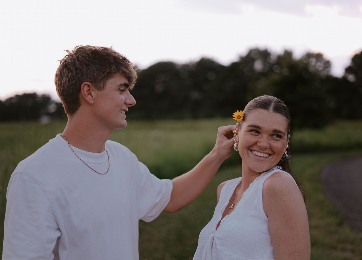 A young man places a yellow flower in a smiling woman's hair outdoors at dusk, both wearing white.