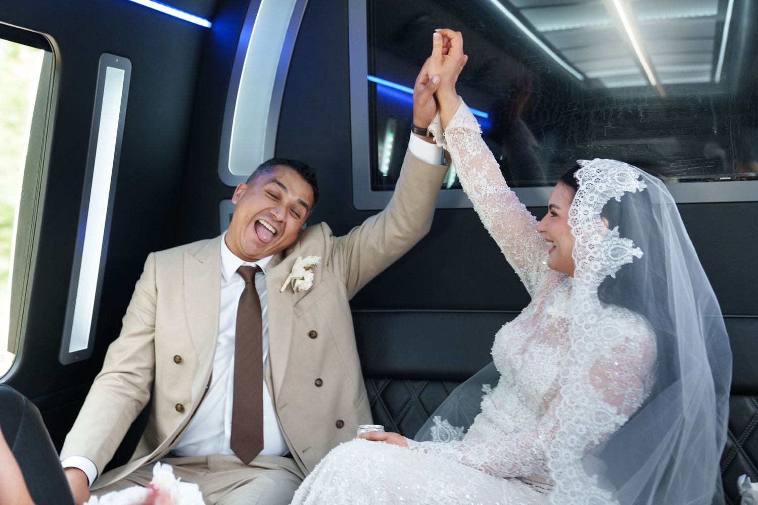 Joyful newlyweds raising hands in celebration inside a limousine, bride in white lace veil and groom in beige suit.