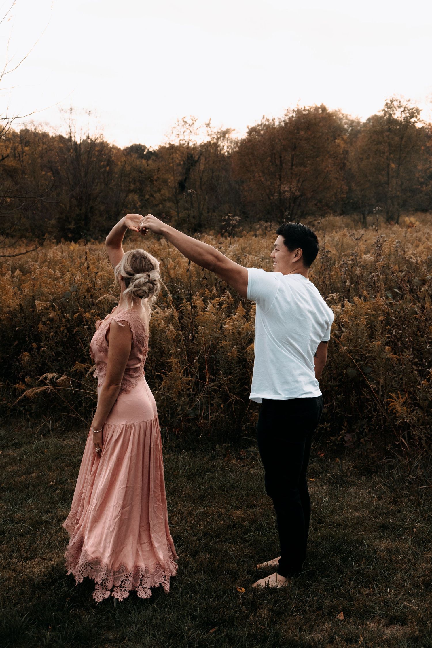 A couple dances outdoors in an autumn field, woman in pink lace dress being twirled by man in white shirt.