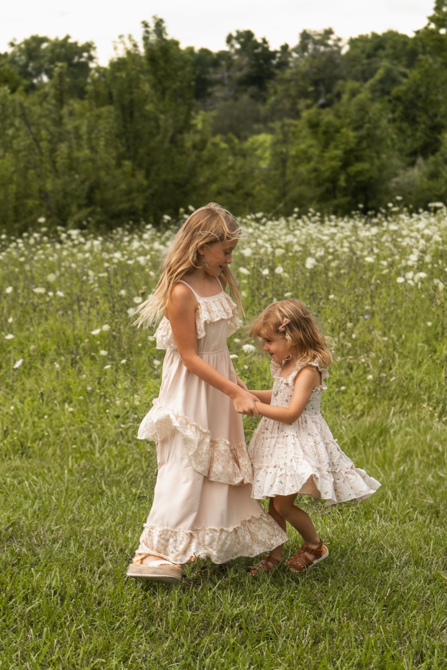 Two figures in matching white flowy dresses dance together in a summer meadow filled with wildflowers.