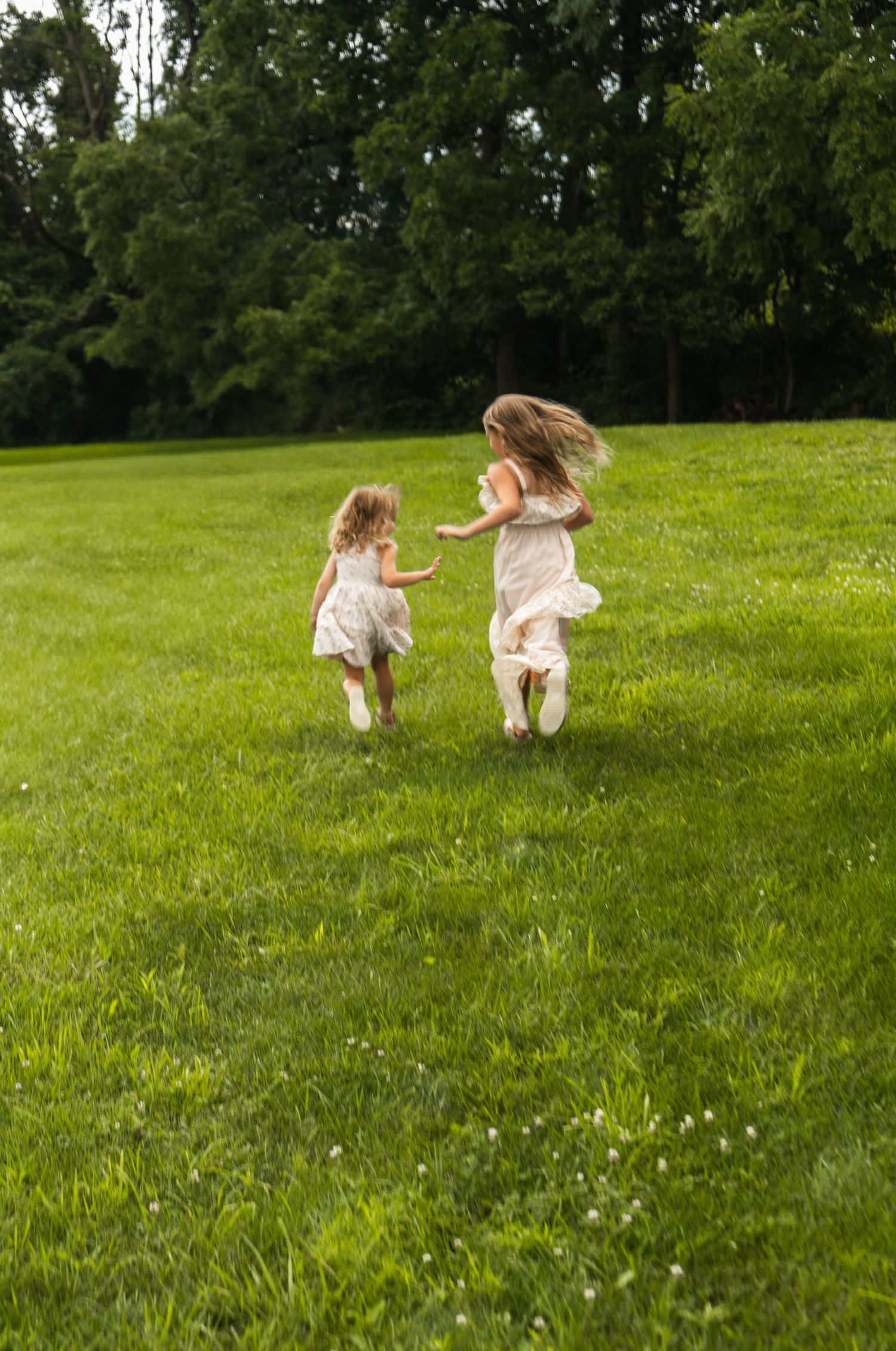 Two young children run and play together on a lush green hillside dotted with wildflowers on a summer day.