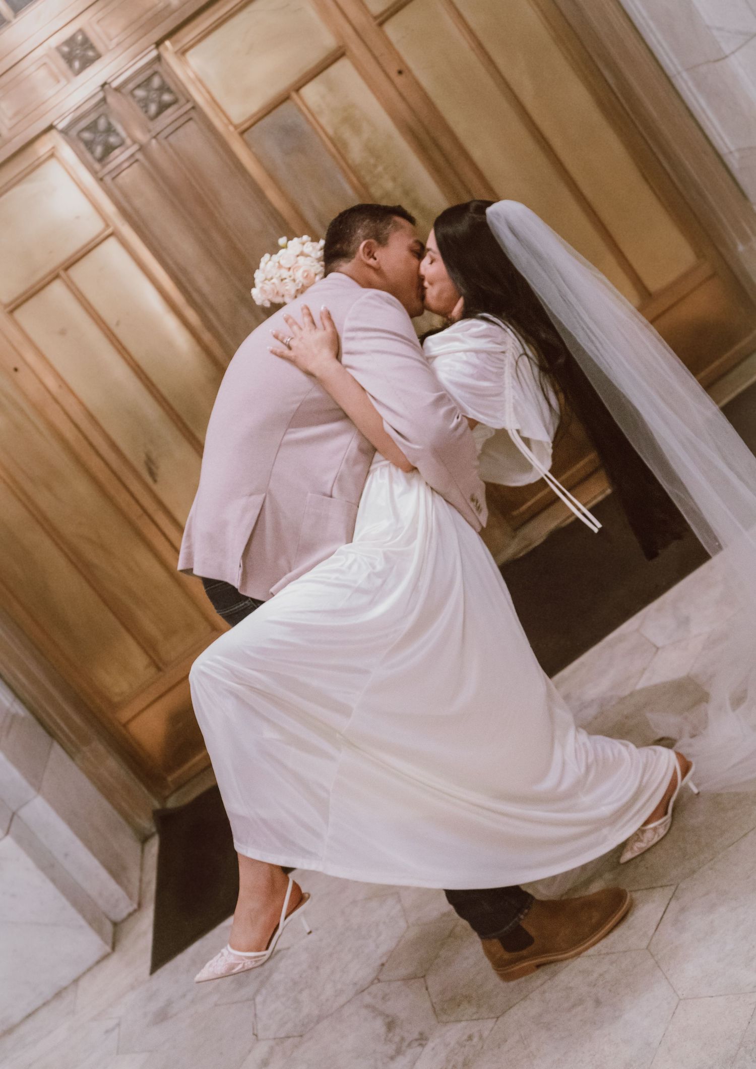 A romantic wedding couple shares an intimate dip pose in a rustic wooden hallway wearing white and pink attire.