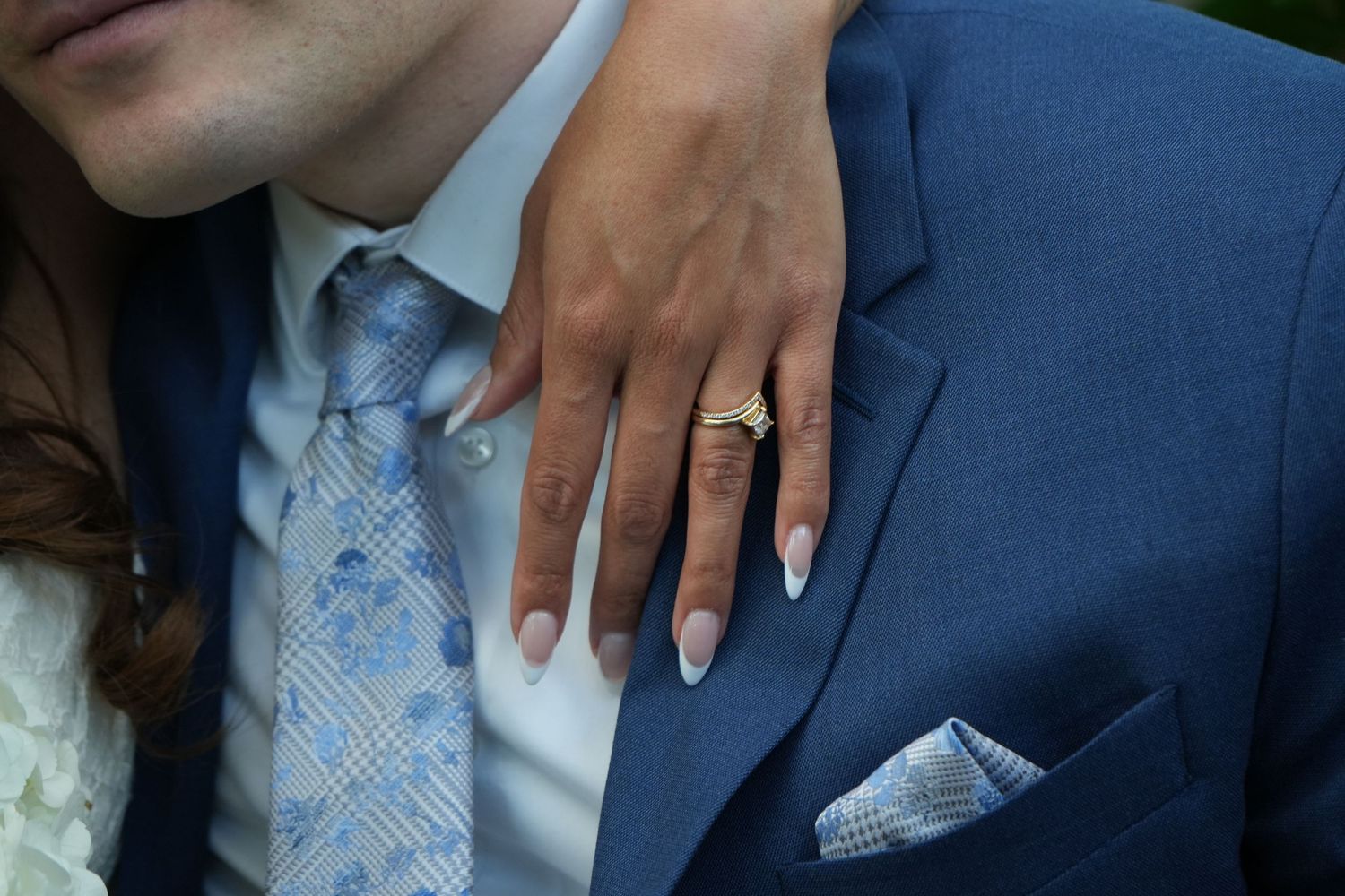 Close-up of a wedding ring displayed on a hand against a blue suit with matching tie and pocket square.